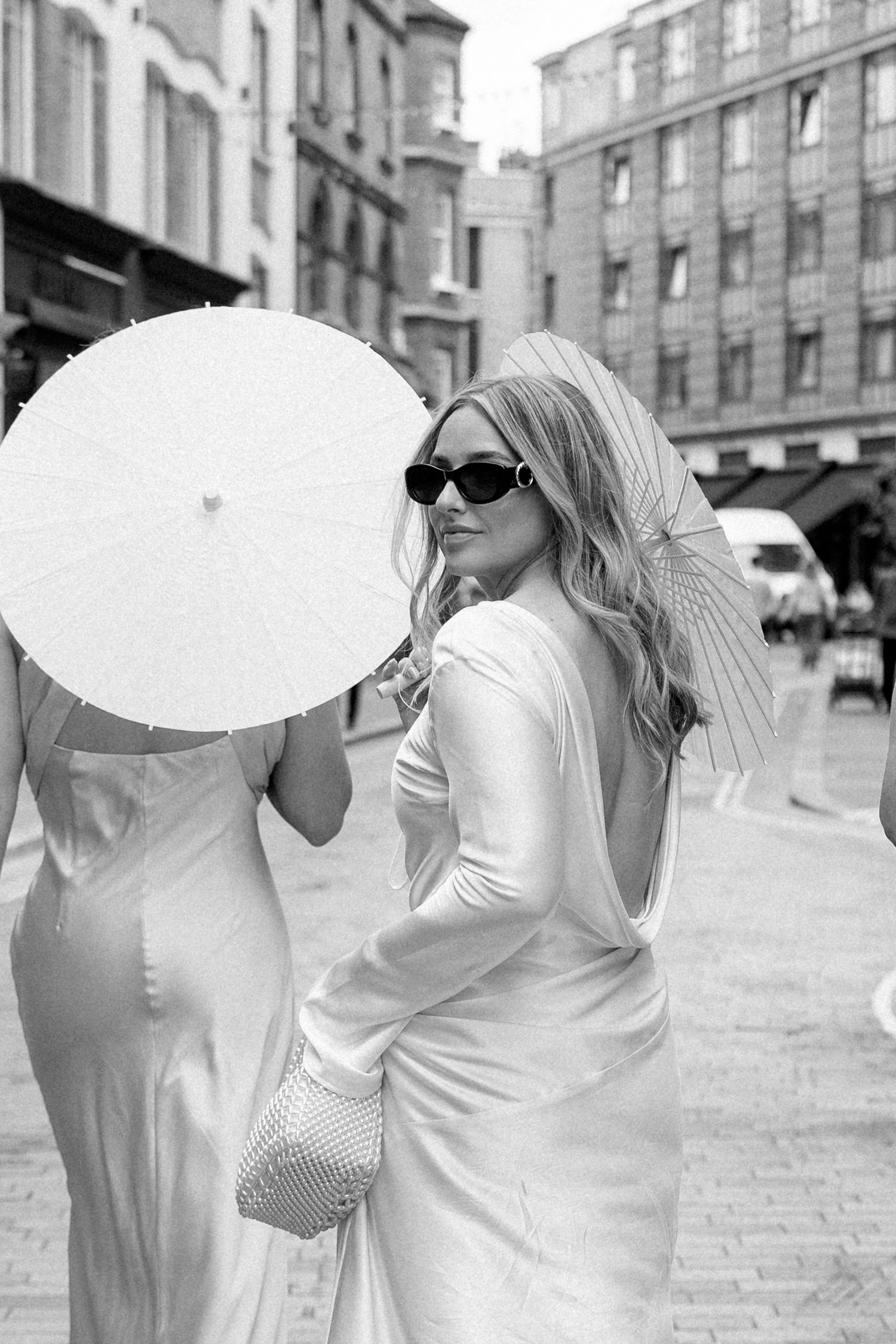 Wedding party members in white dresses carry parasols on a city street.