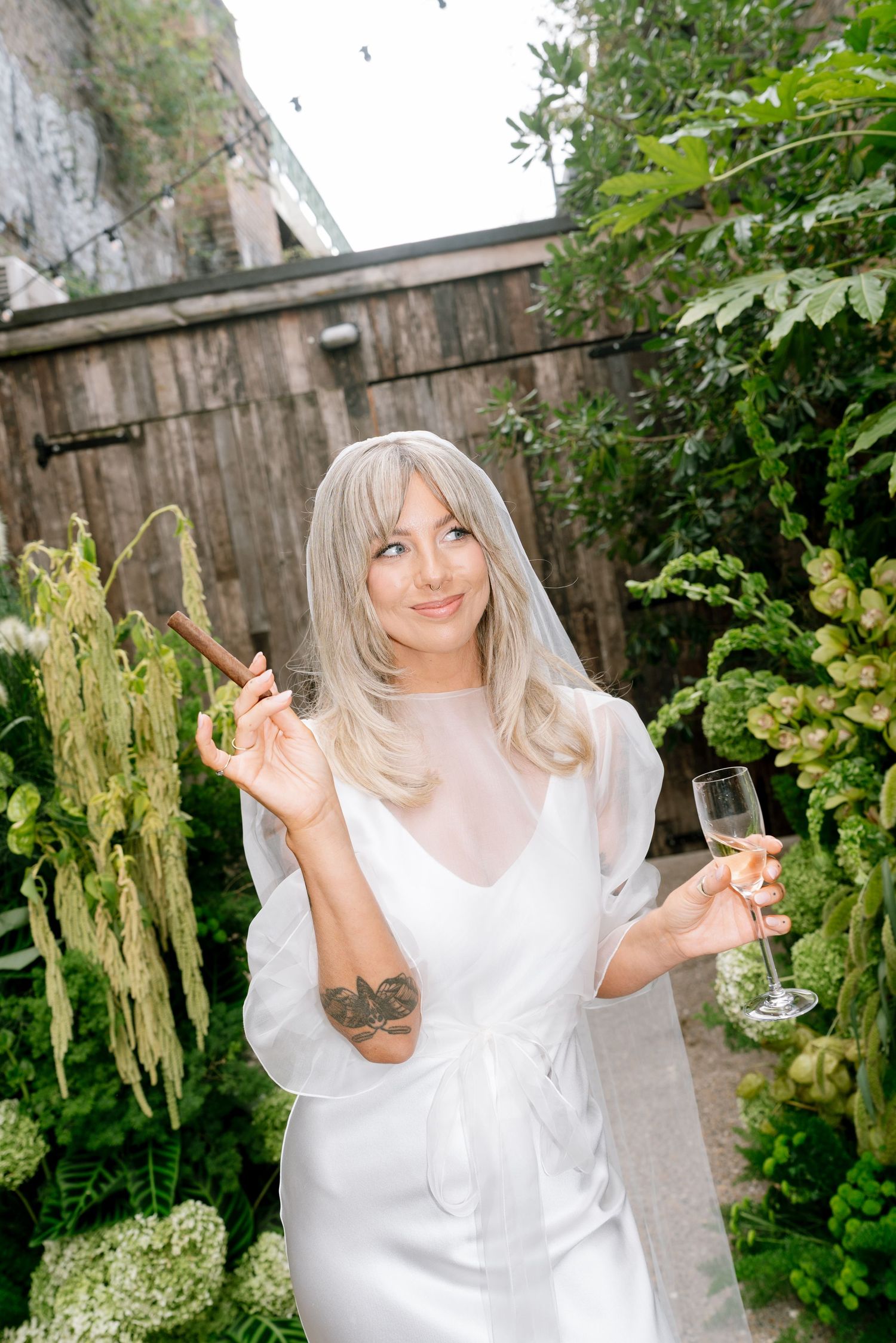 Person in white dress poses playfully in a garden setting with a drink.