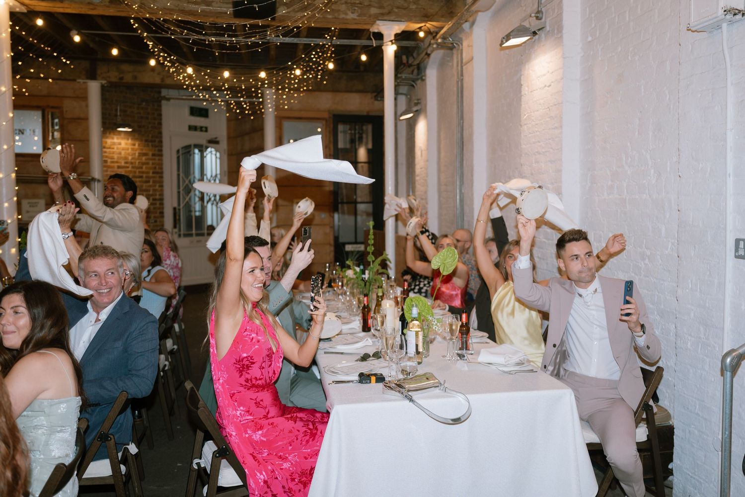 Wedding reception guests raise their glasses in celebration under string lights at an indoor venue.