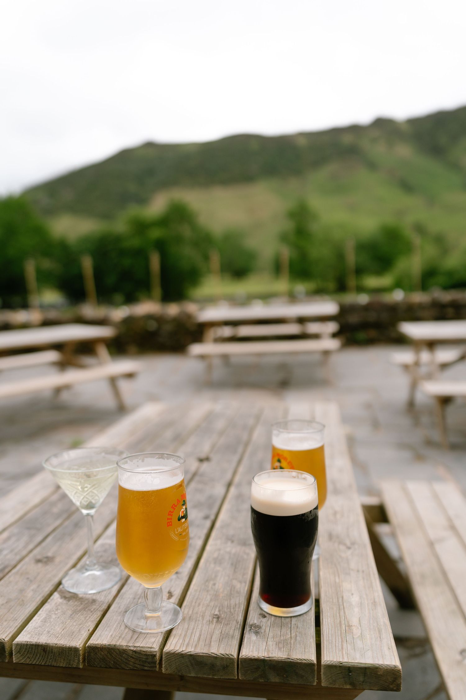 Three craft beer glasses with varying colors from light to dark sit on a wooden picnic table with mountains in background.