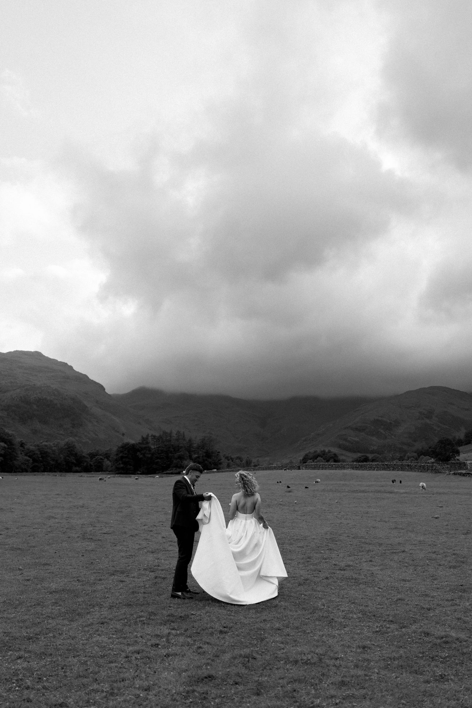 Wedding couple poses in a scenic field with dramatic cloudy mountains as backdrop.