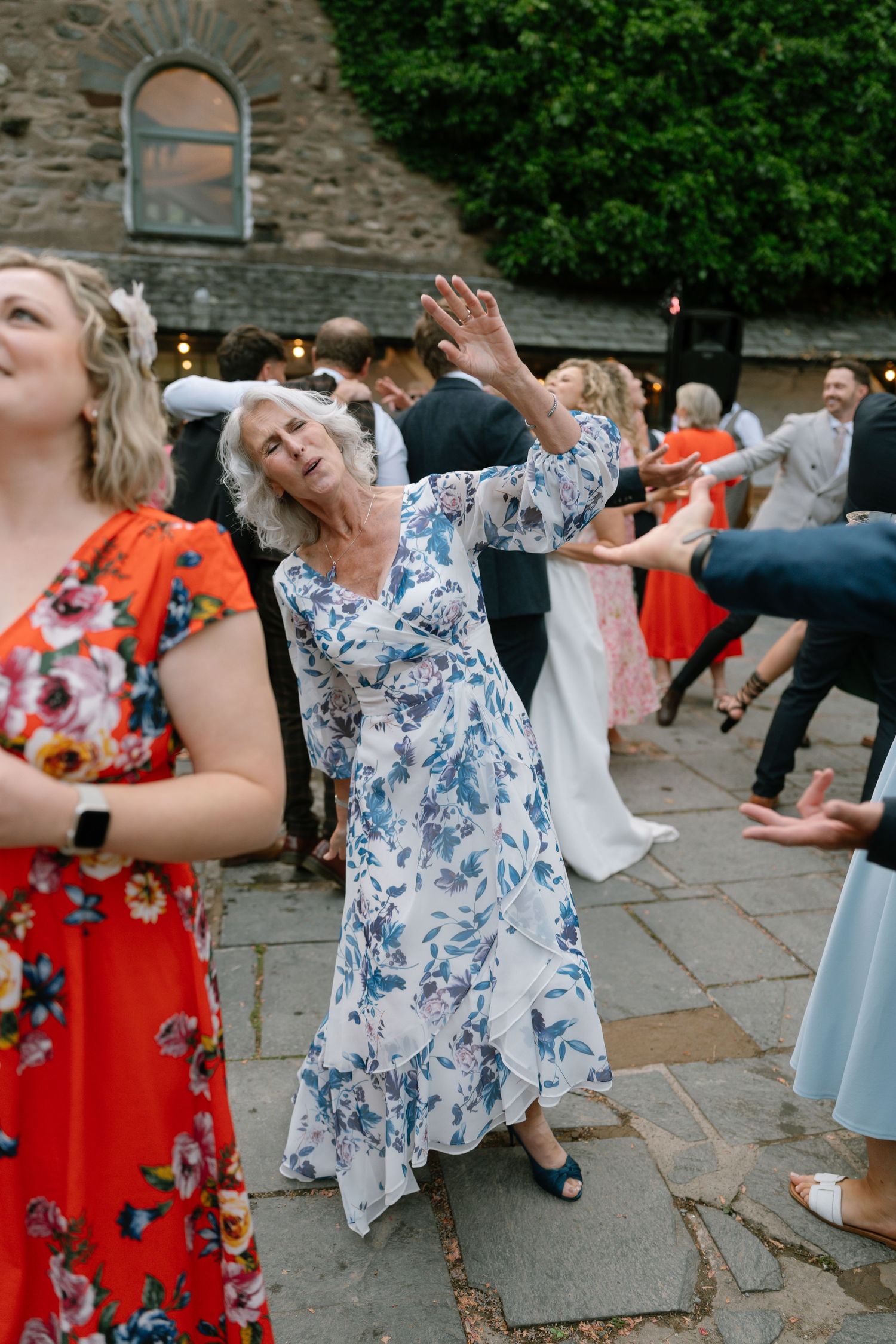 Wedding guests dancing and celebrating on a stone patio near an old stone building with arched window.