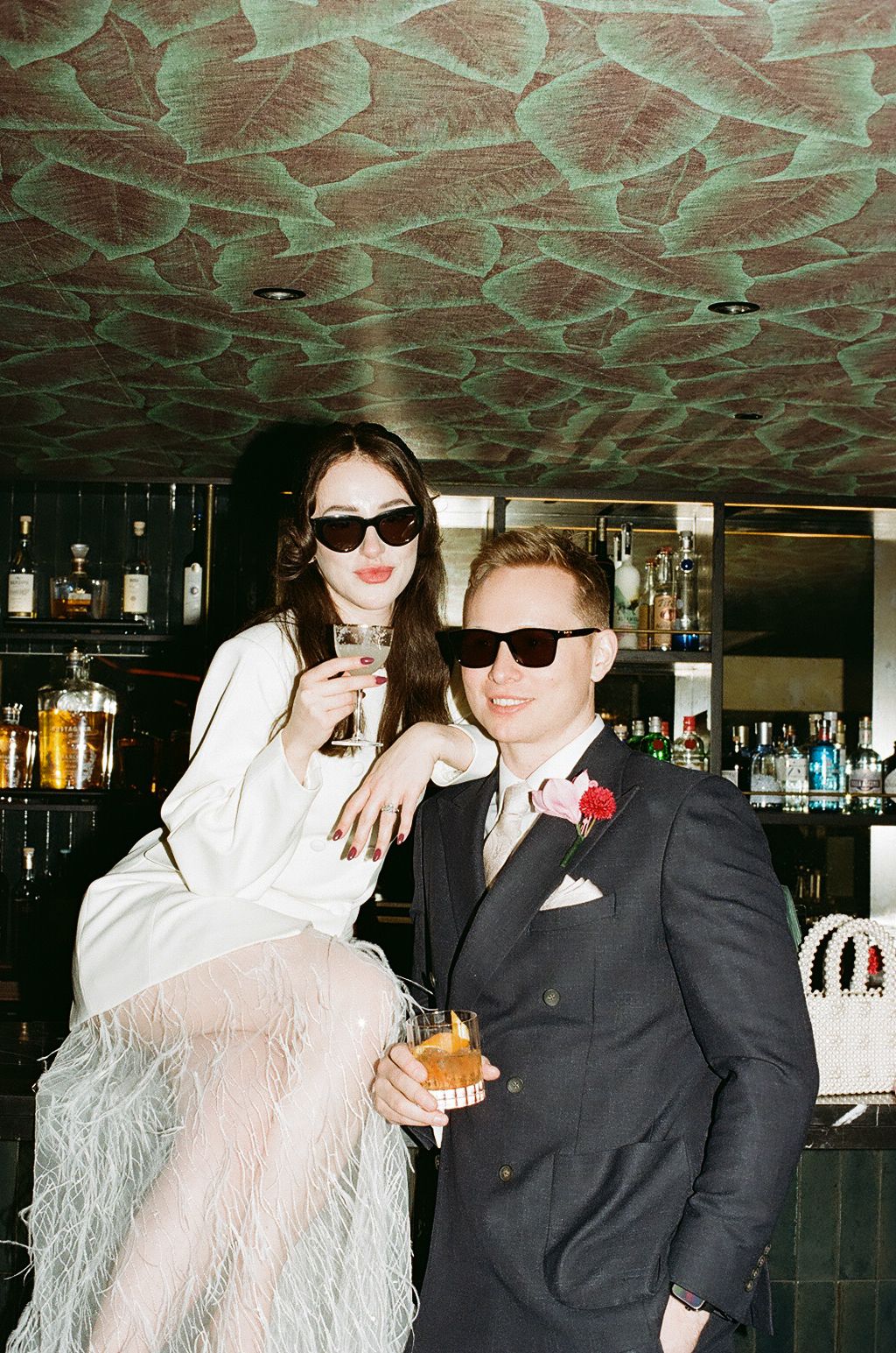Two people in sunglasses pose at a bar with drinks, one wearing a white feathered outfit and one in a dark suit.