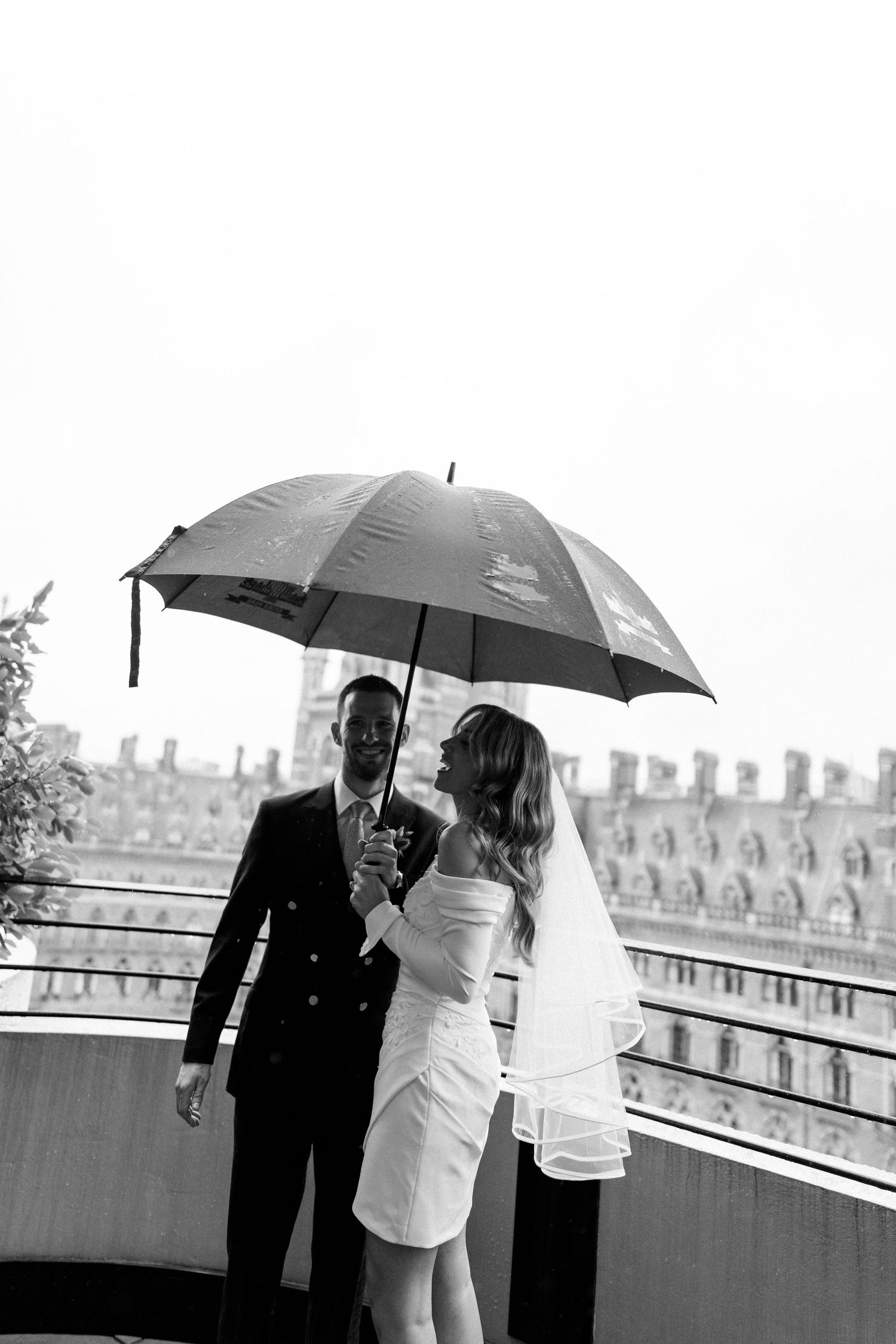 A couple shares a romantic moment under a black umbrella on a rainy day in Paris, with historic buildings in the background.
