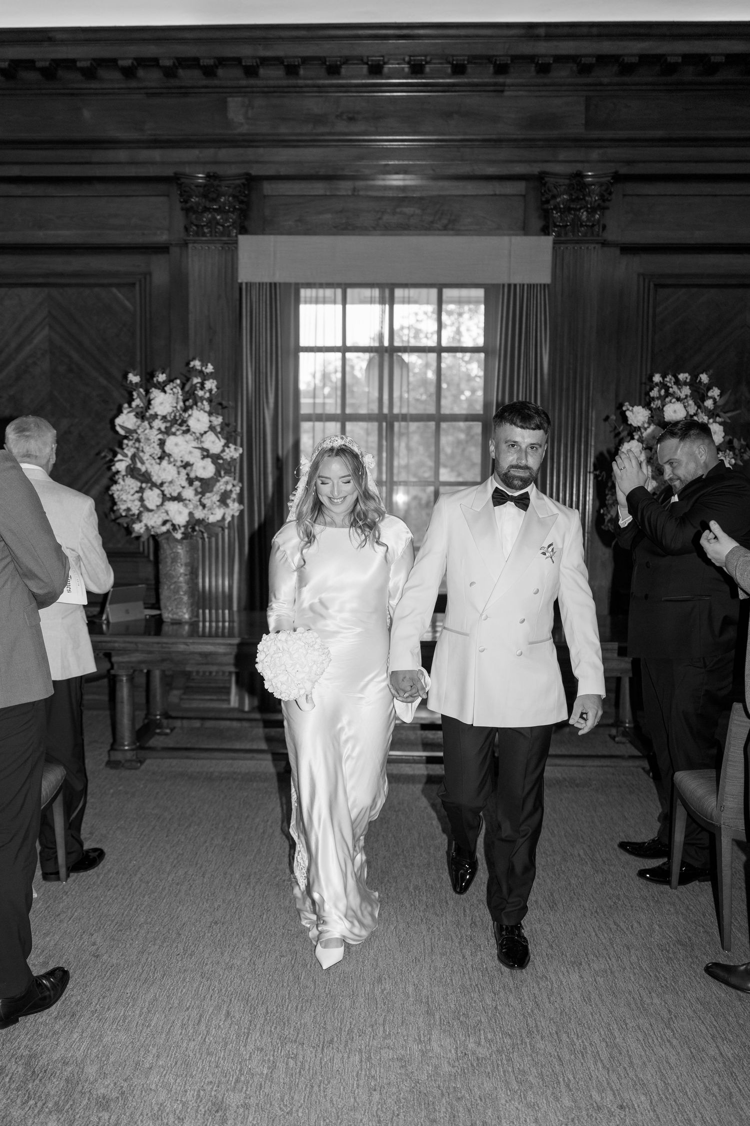 Black and white photo of newlyweds walking down aisle while guests stand in wood-paneled room.