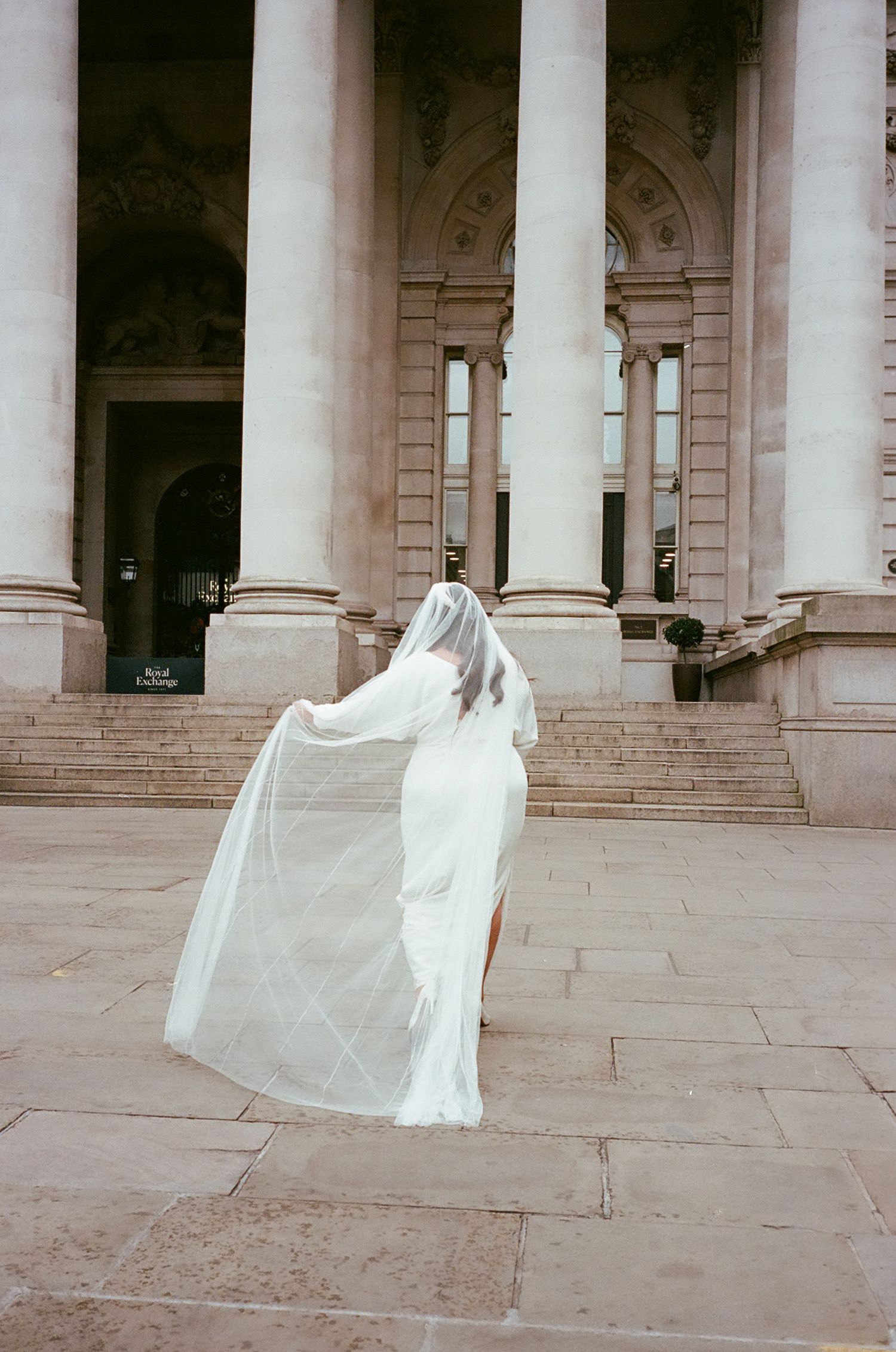 A flowing white wedding dress and veil captured in motion on grand stone steps with classical columns in the background.