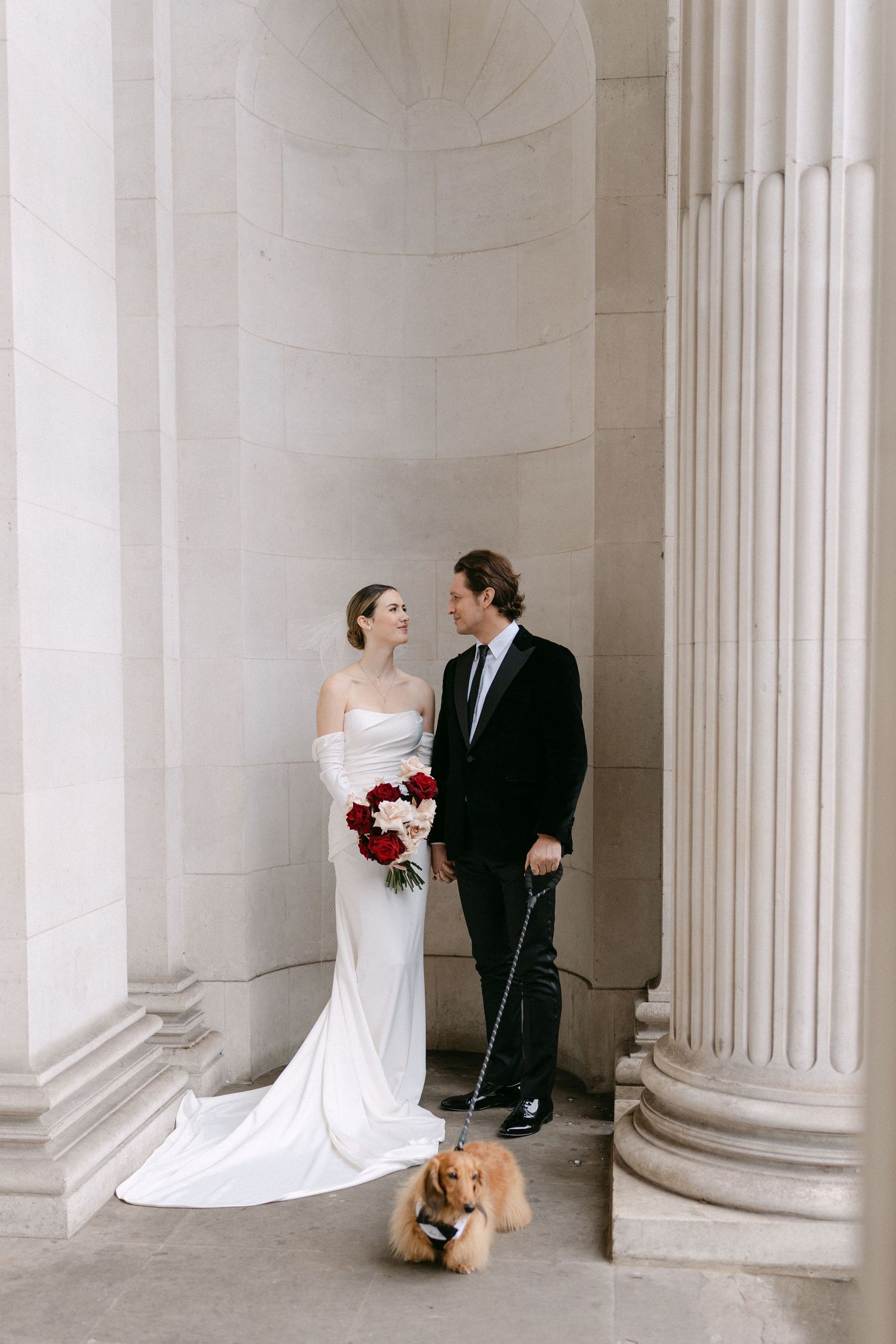 A wedding couple poses between classic white columns in an elegant architectural setting.