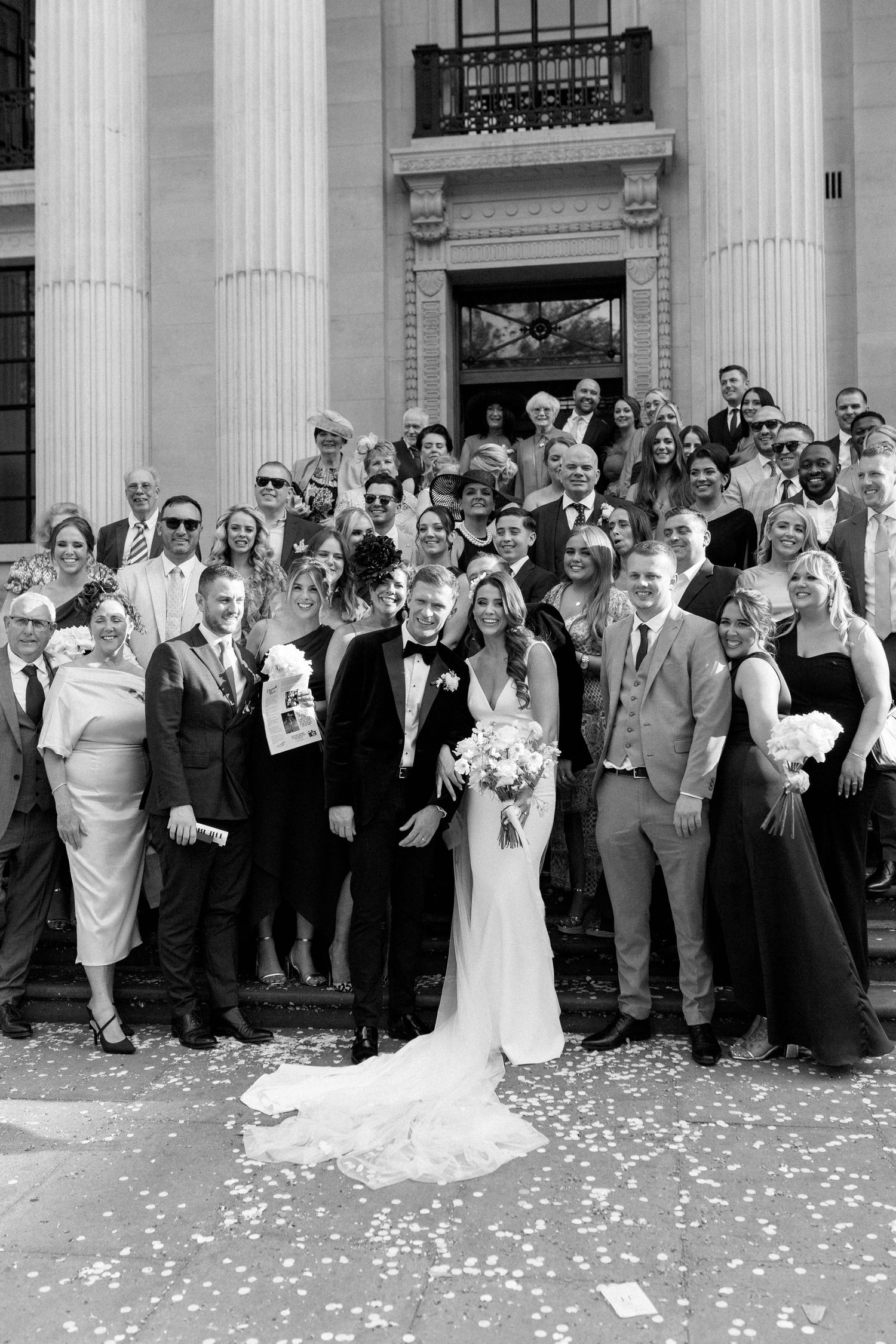 Large wedding party poses for formal group photo on historic building steps with flower petals.