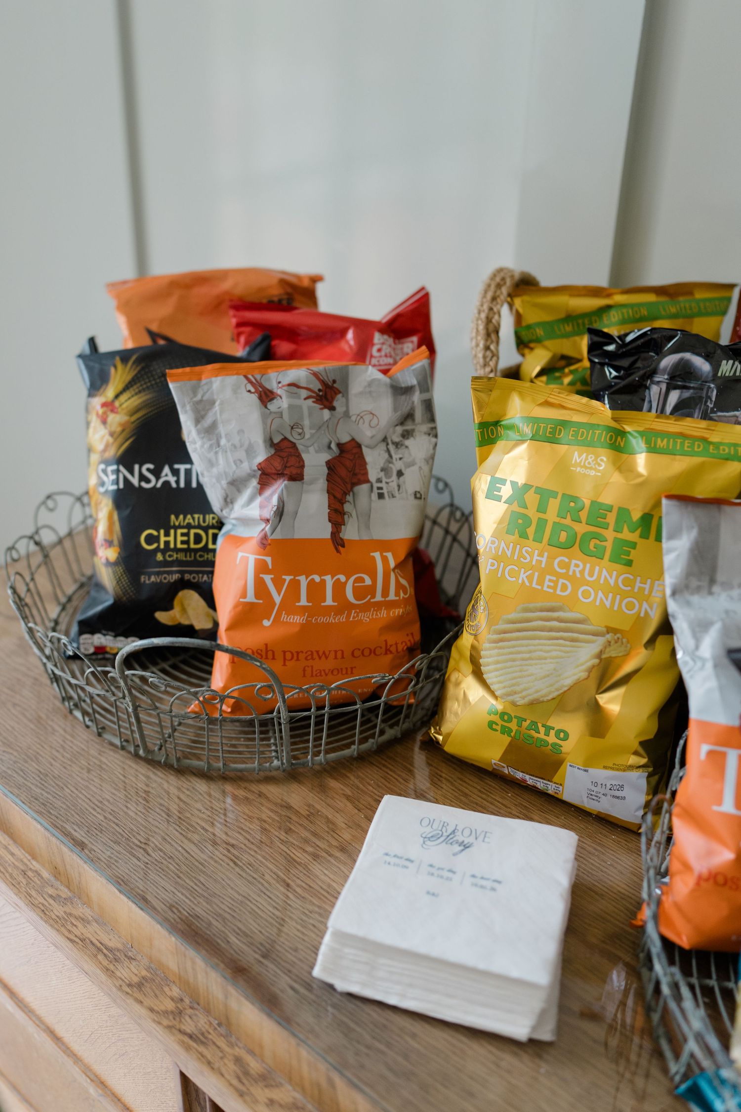 Wire basket filled with Tyrrells, Sensations, and Extreme Ridge crisp packets on a wooden table with napkins.