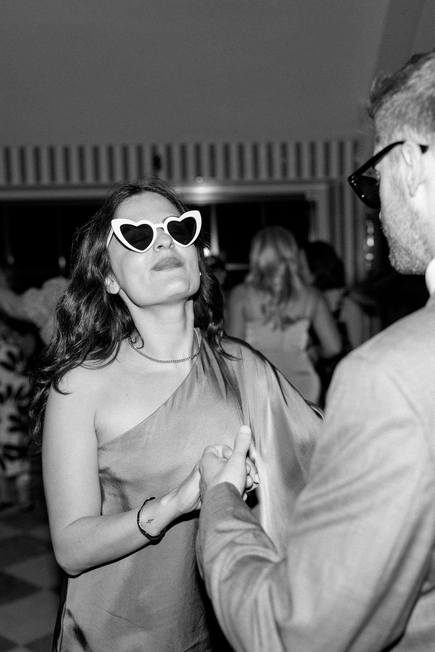 Black and white photo of wedding guest dancing in heart-shaped sunglasses at evening reception.