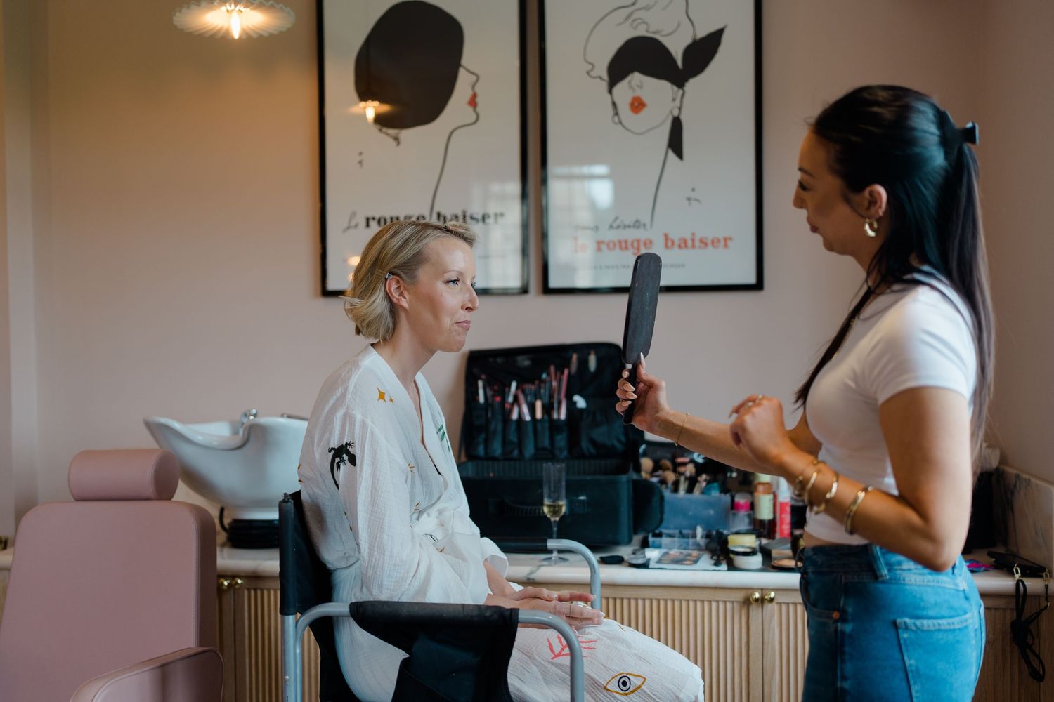 Bride in white robe consulting with hairstylist holding mirror in elegant pink wedding preparation salon.