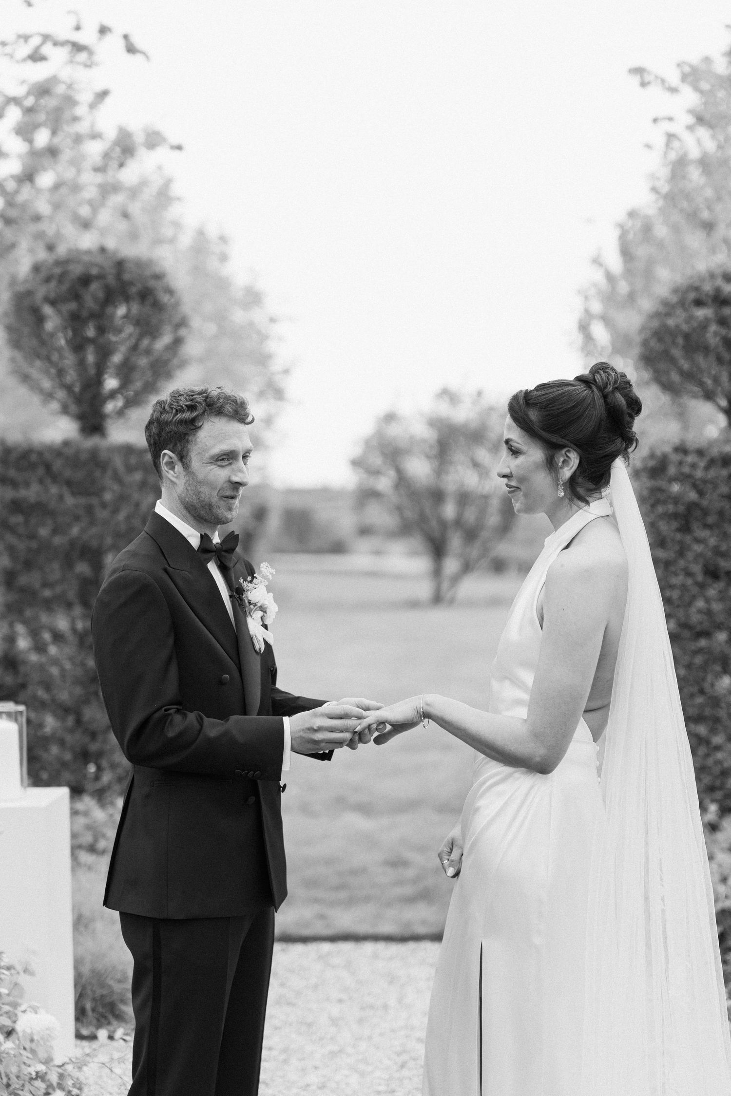Black and white photo of bride and groom exchanging rings during outdoor garden wedding ceremony.