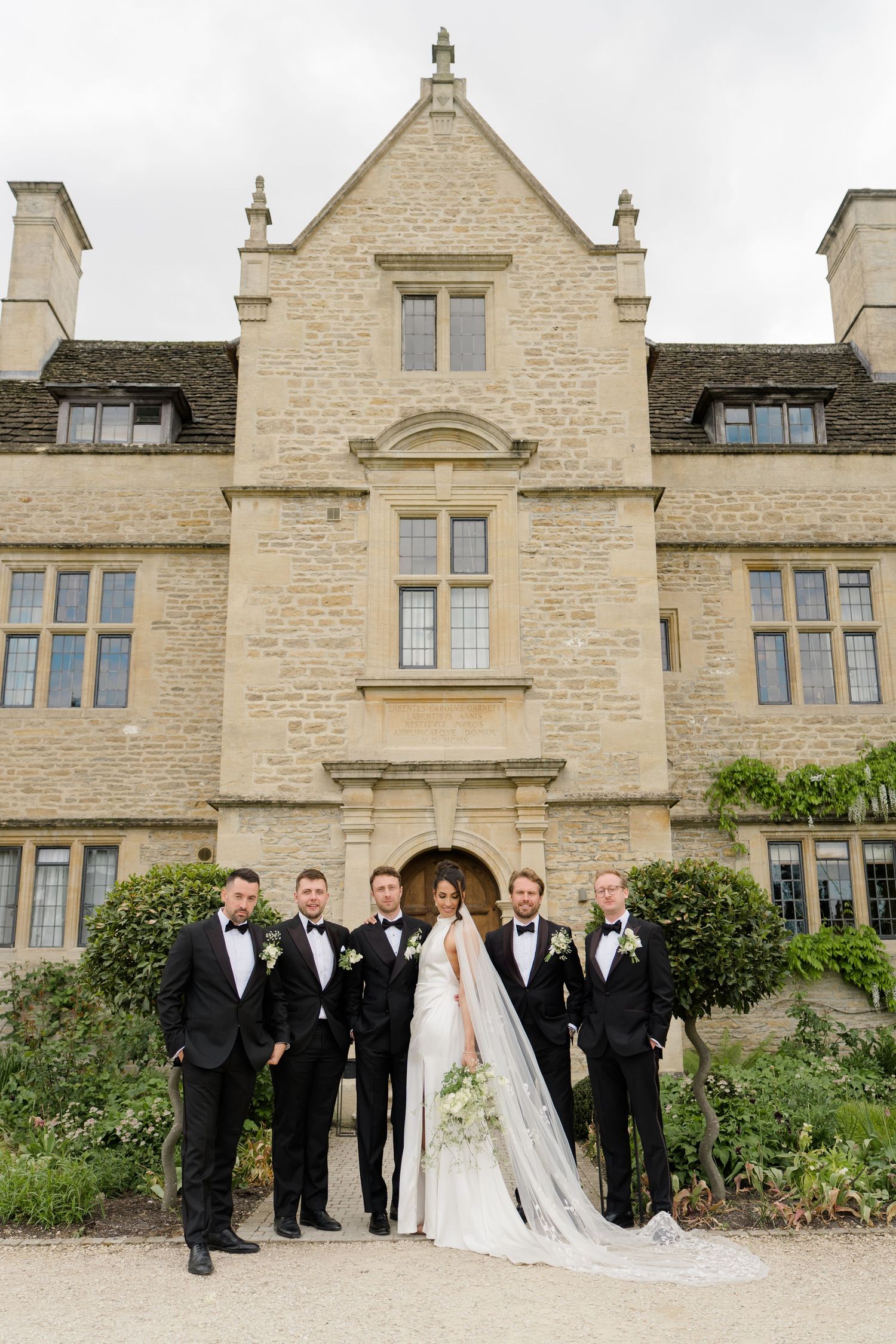 Bride and groomsmen in black tuxedos pose formally before a grand stone English manor house.