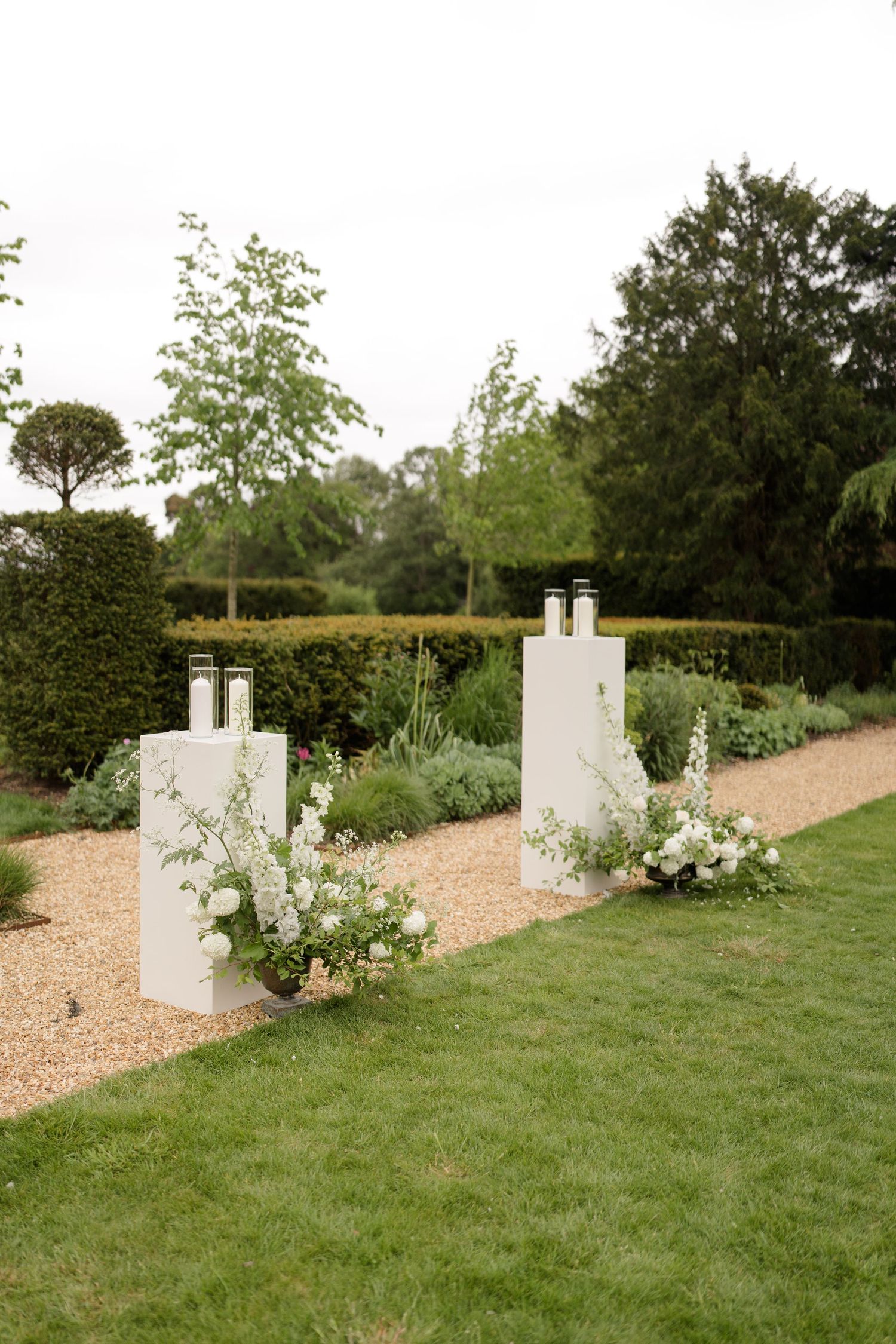 White pedestals with lanterns and floral arrangements flanking a garden path at an outdoor wedding ceremony.