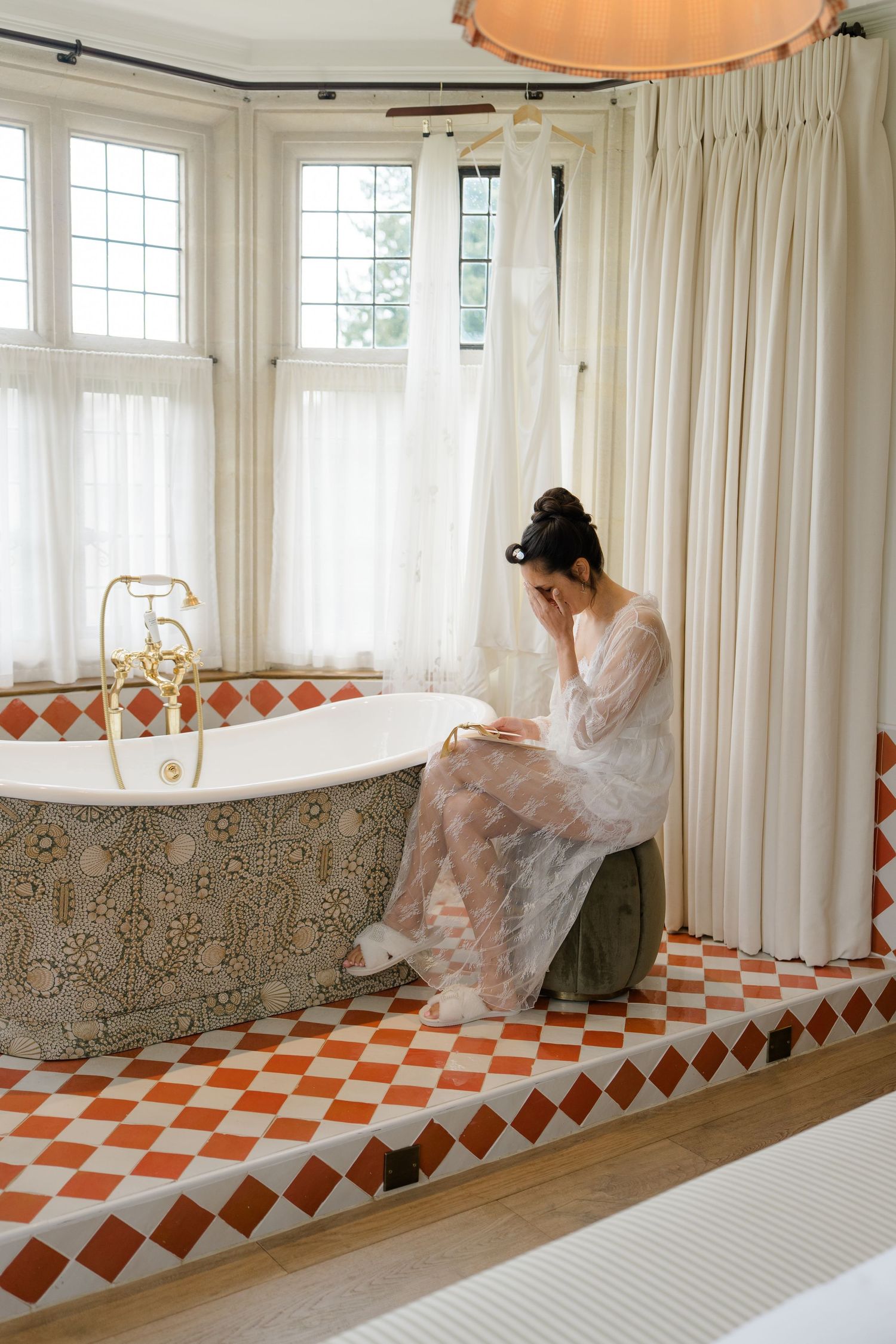 Bride in lace robe sitting beside ornate freestanding bathtub on red checkered tile floor.