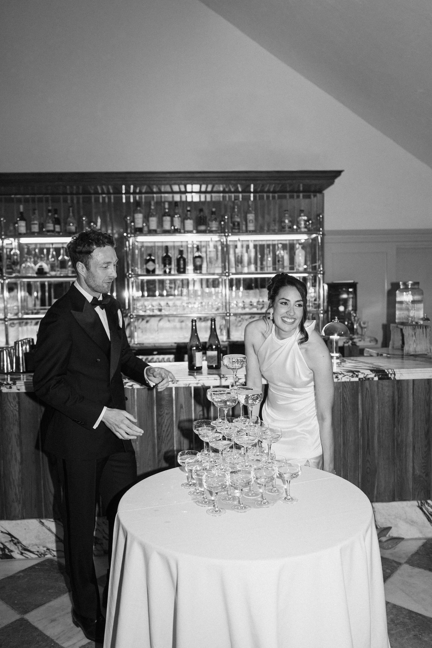 Bride and groom in black and white photo standing beside champagne tower at wedding bar.