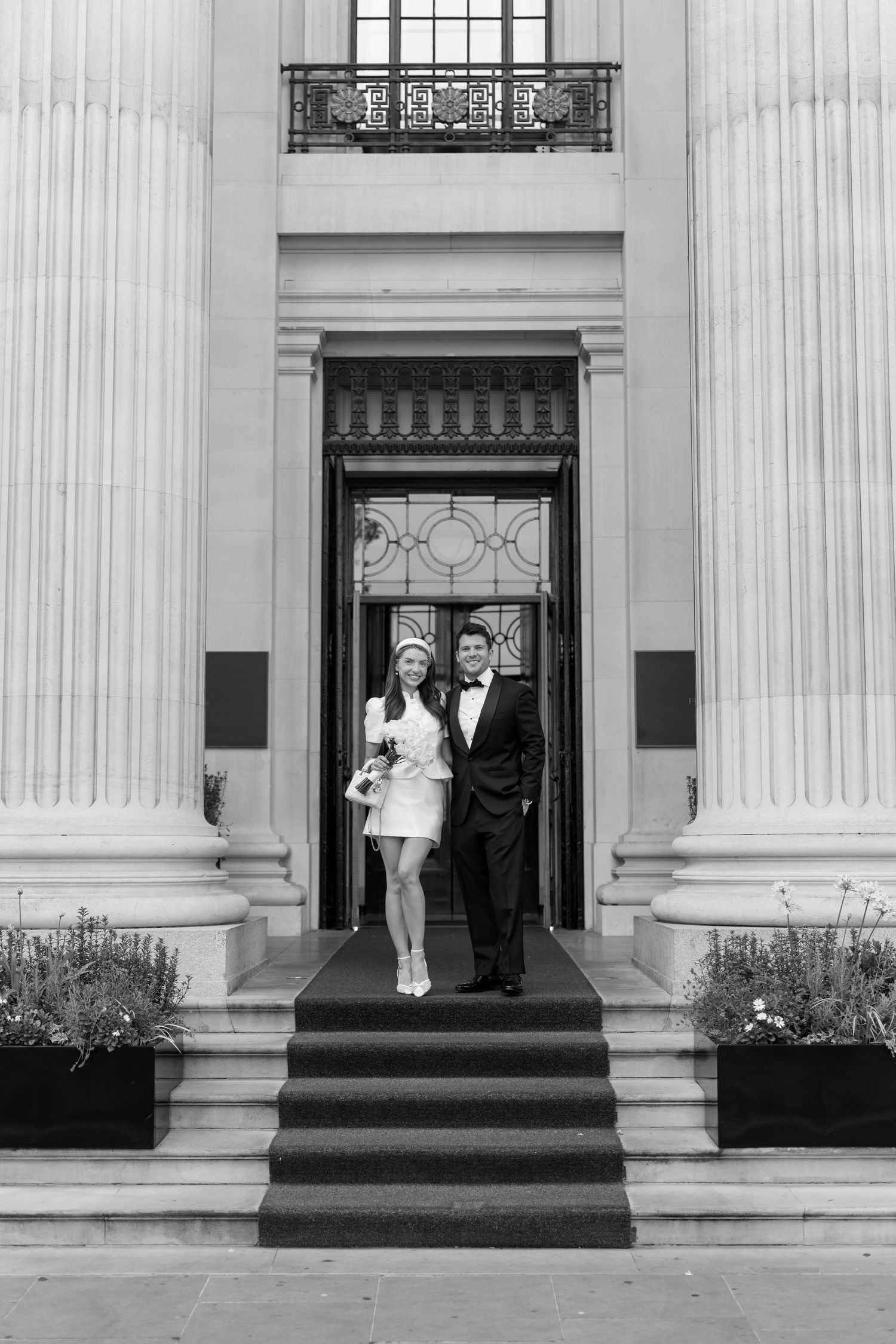 Elegant bride and groom in black tuxedo descending grand building steps in black and white wedding photo.
