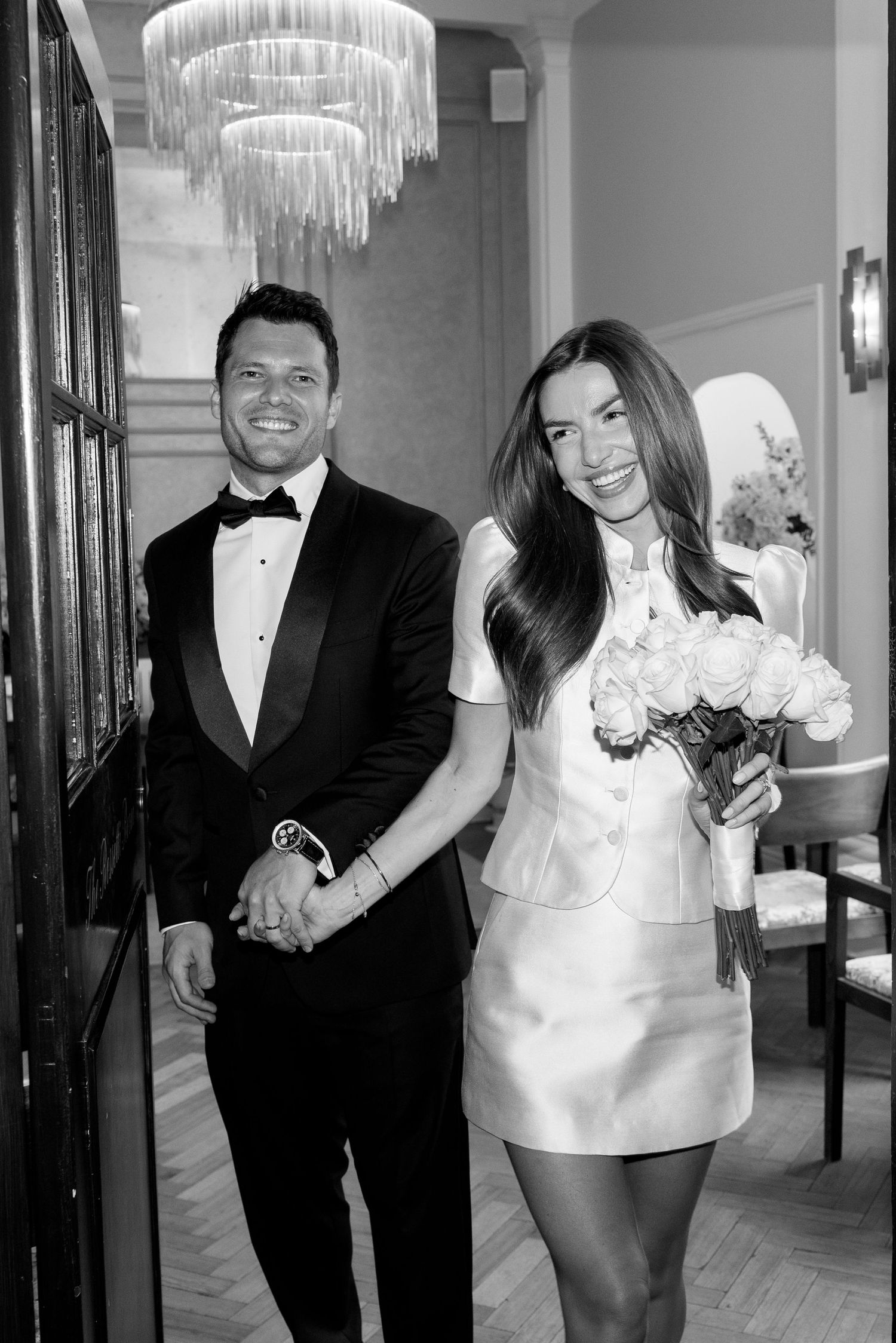 Black and white portrait of smiling bride and groom in tuxedo holding white rose bouquet in elegant interior.