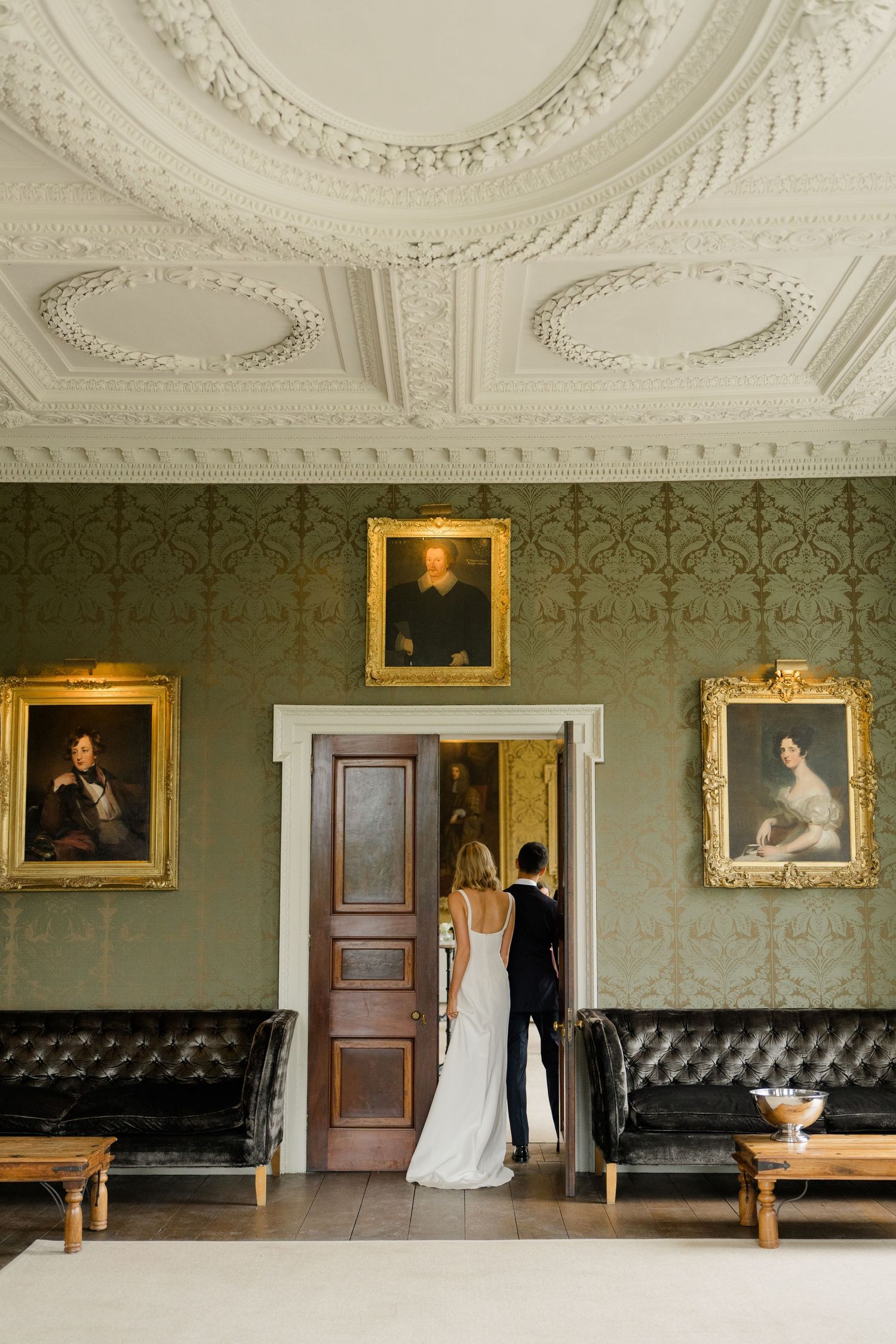 Bride and groom stand in ornate room with portrait paintings and decorative plasterwork ceiling.