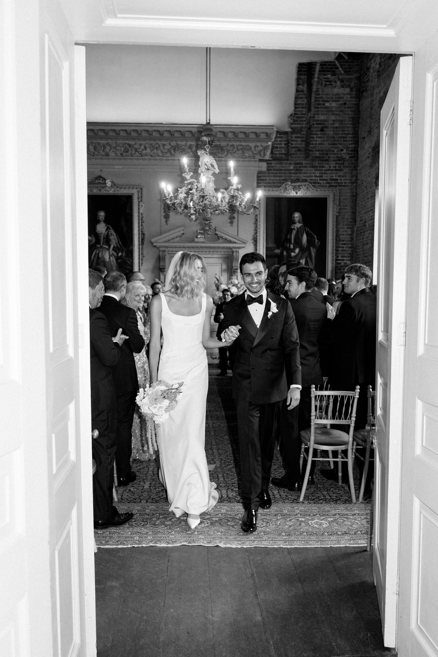 Bride and groom walk down the aisle together in a grand chandelier-lit hall surrounded by wedding guests.