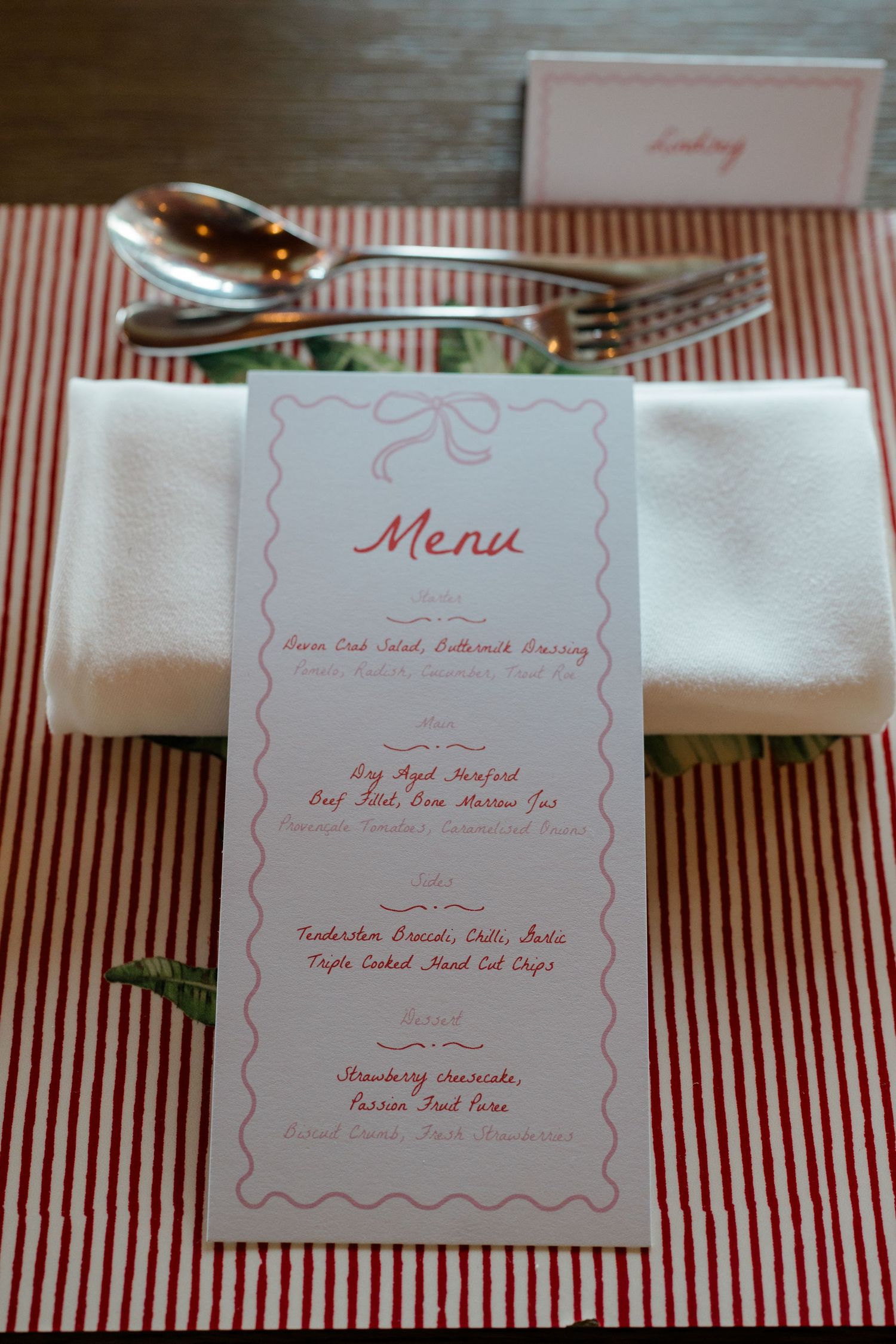 Elegant wedding place setting with menu card, white napkin, and silverware on red striped tablecloth.