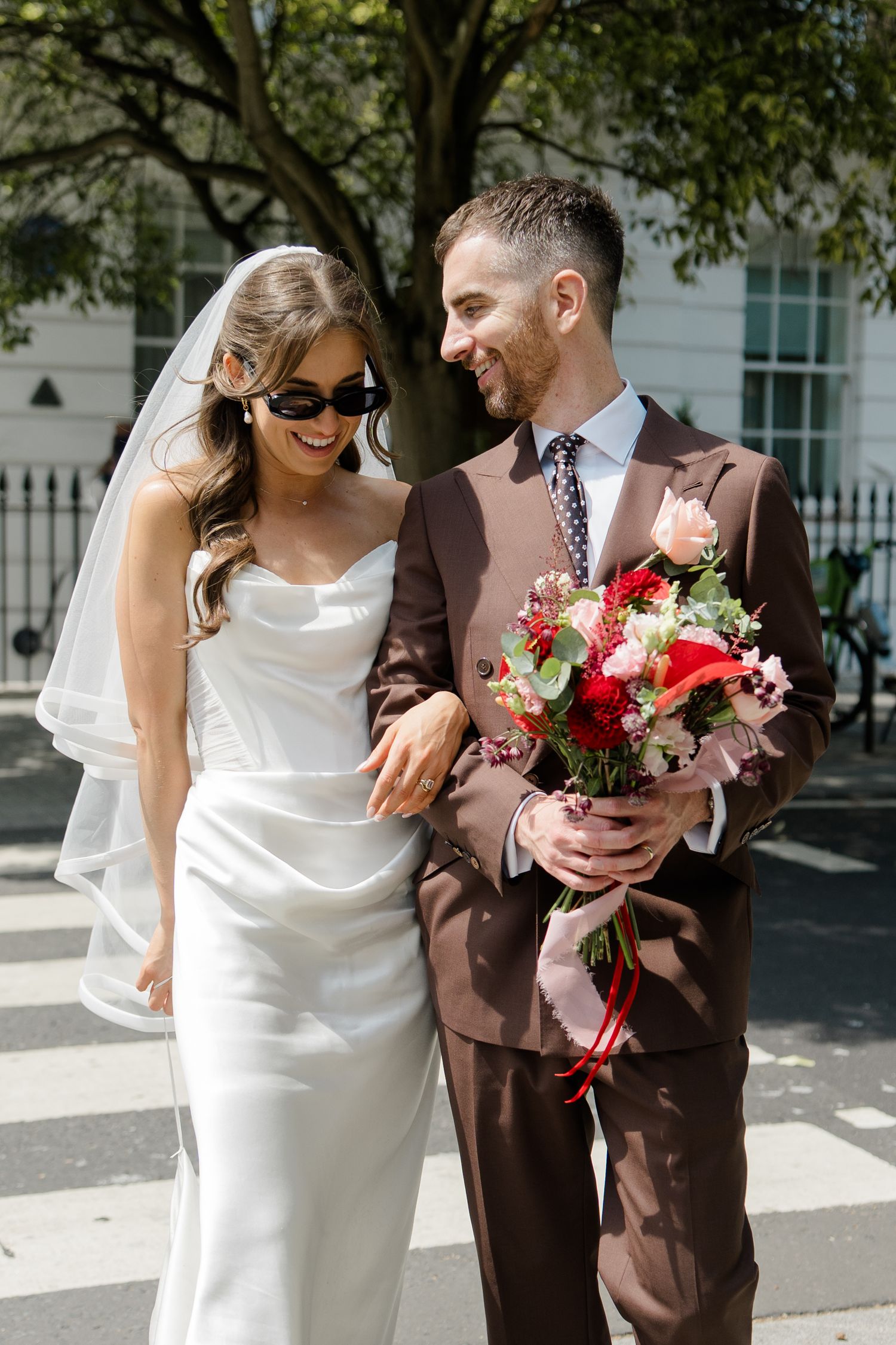 Happy bride and groom in sunglasses smile together on a sunny street, groom holding bouquet.