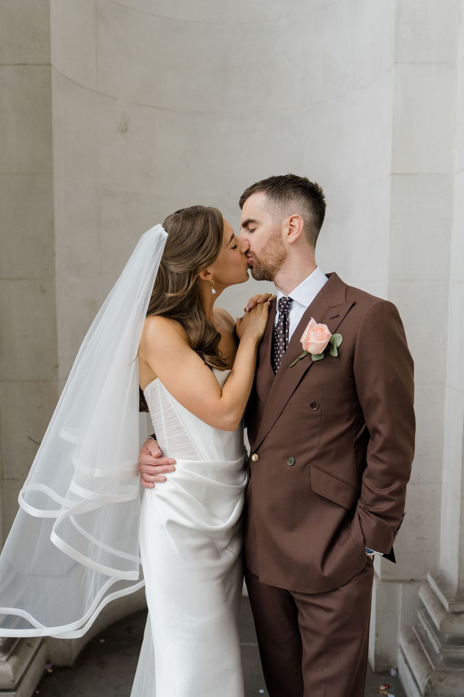Bride in white gown and veil kisses groom in brown suit with pink boutonniere outdoors.