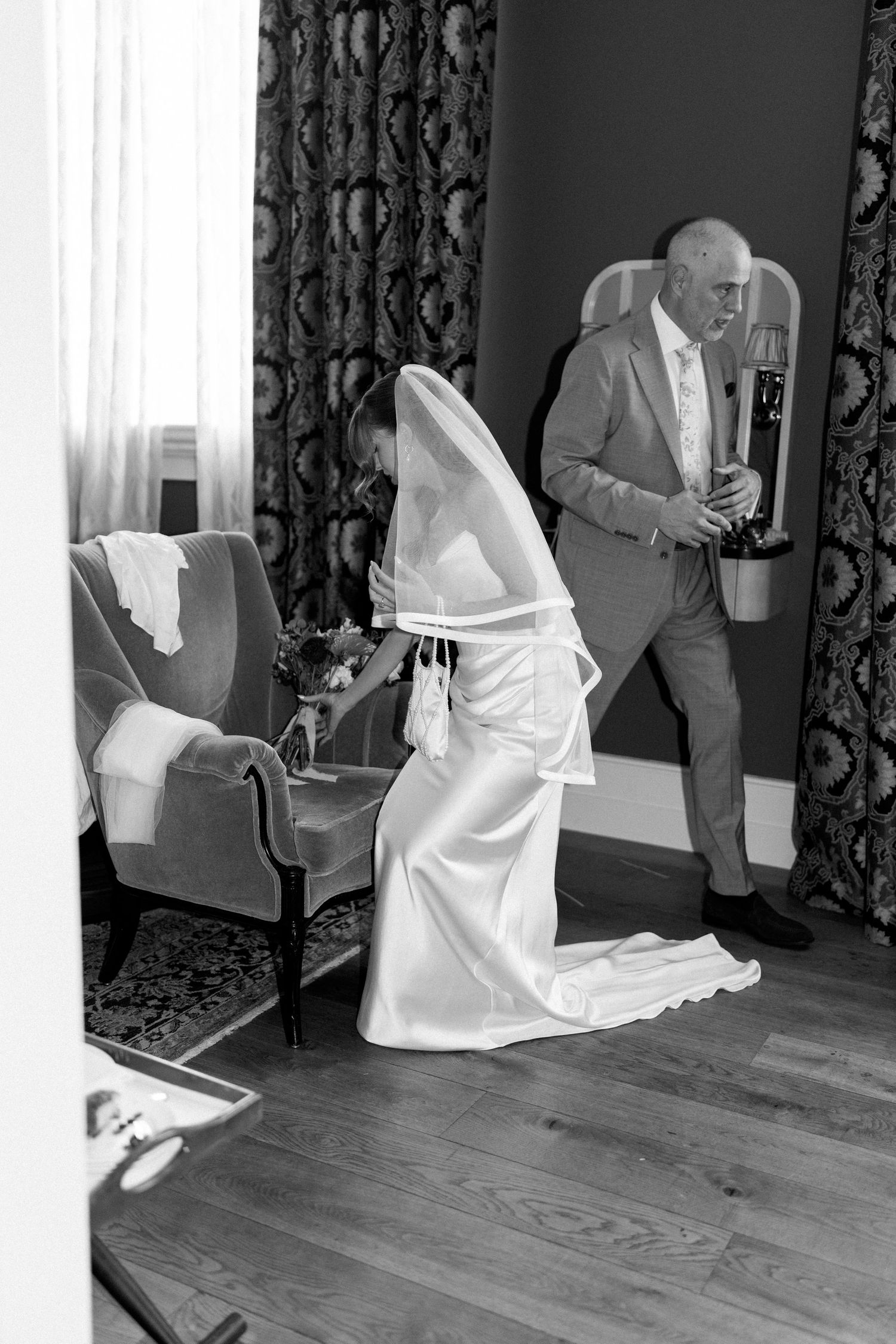 Black and white photo of bride in wedding dress and veil kneeling while man holds champagne bottle.