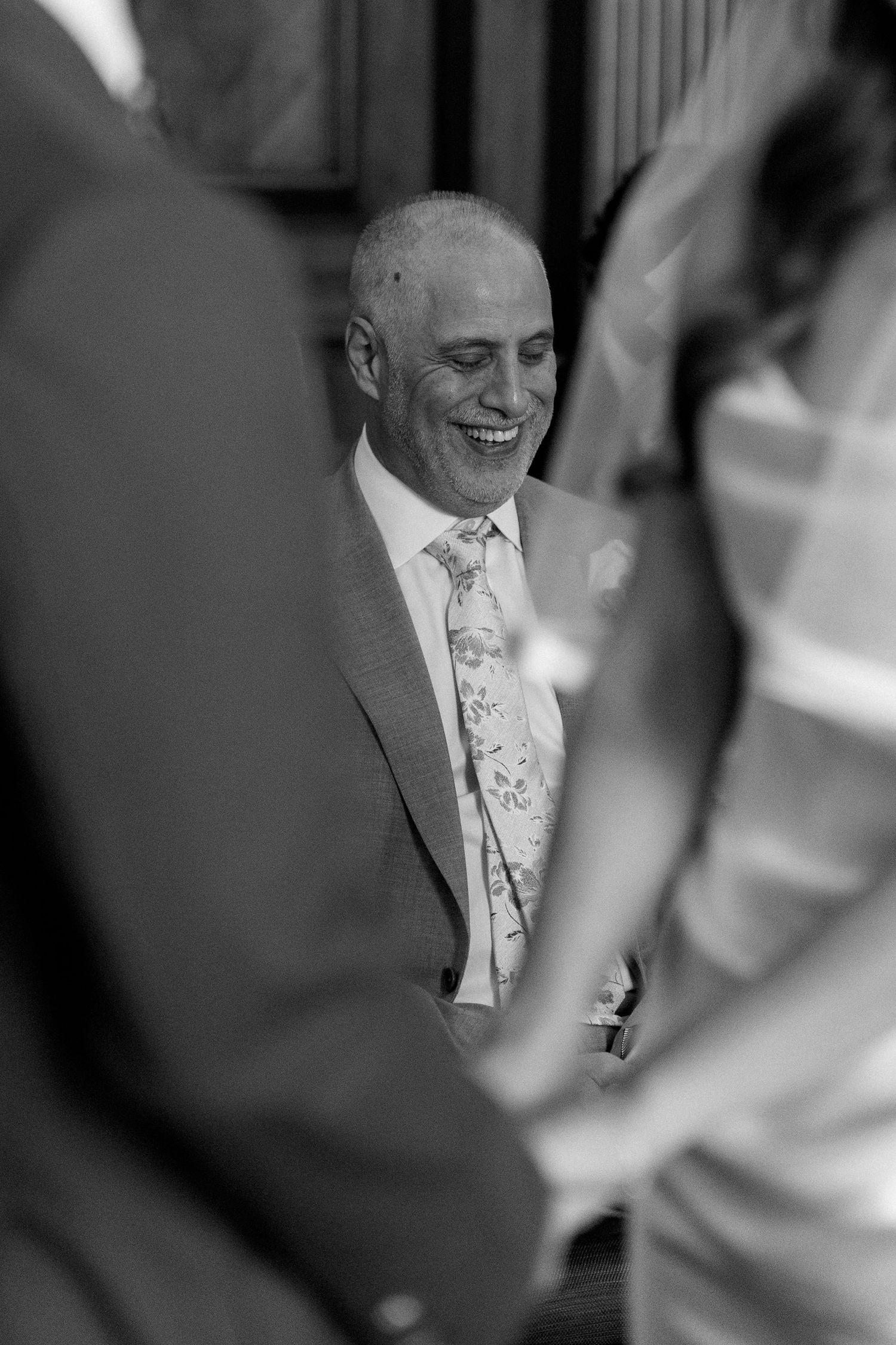 Bald man in grey suit with floral tie laughs joyfully during wedding ceremony in black and white photo.