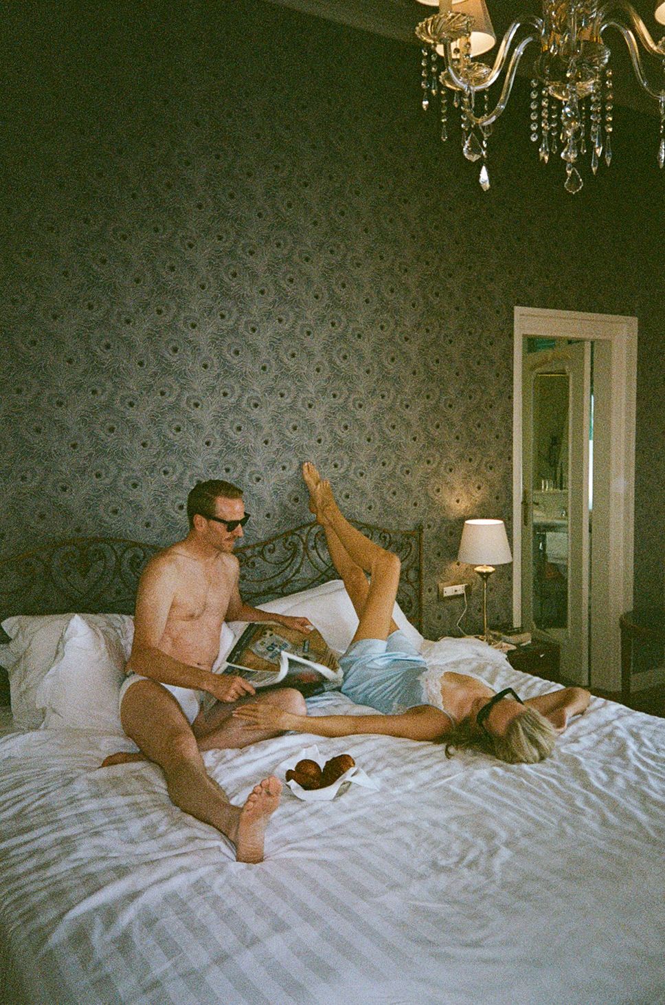 Couple relaxing on a luxurious bed in an elegant room with chandelier and patterned wallpaper.