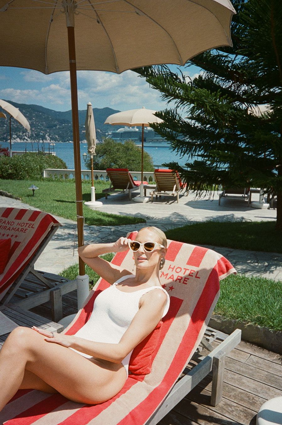 Blonde woman in white swimsuit relaxes on striped Hotel Splendido lounger by the Italian Riviera pool.