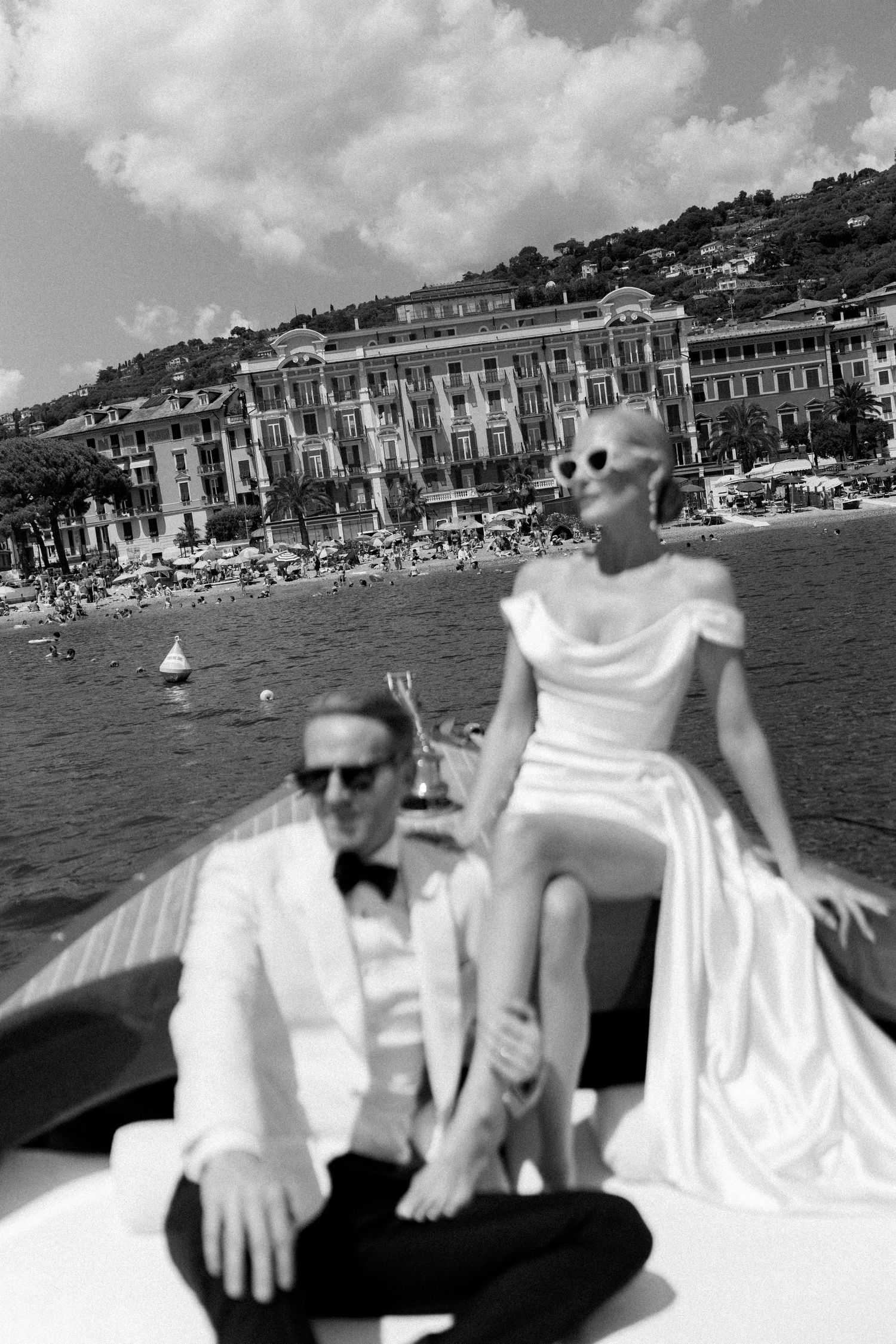 Bride in white gown posing on luxury boat with groom in white tuxedo, Italian coastal town backdrop.