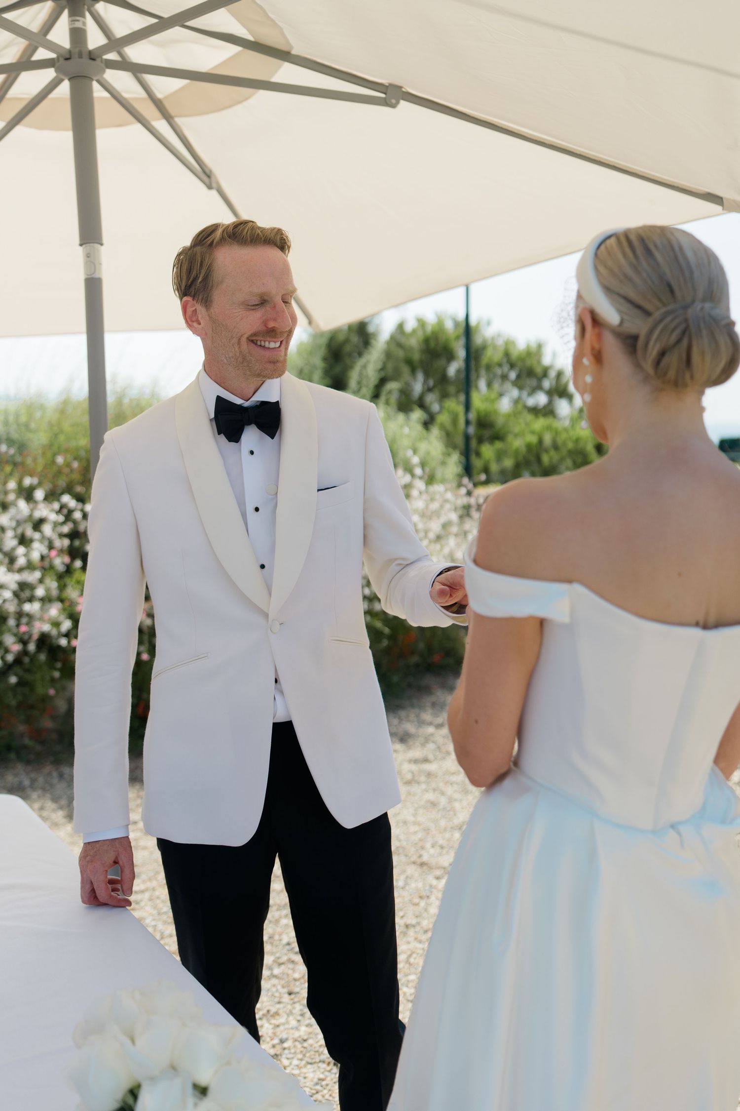 Groom in white tuxedo smiles at bride in off-shoulder dress under outdoor umbrella.