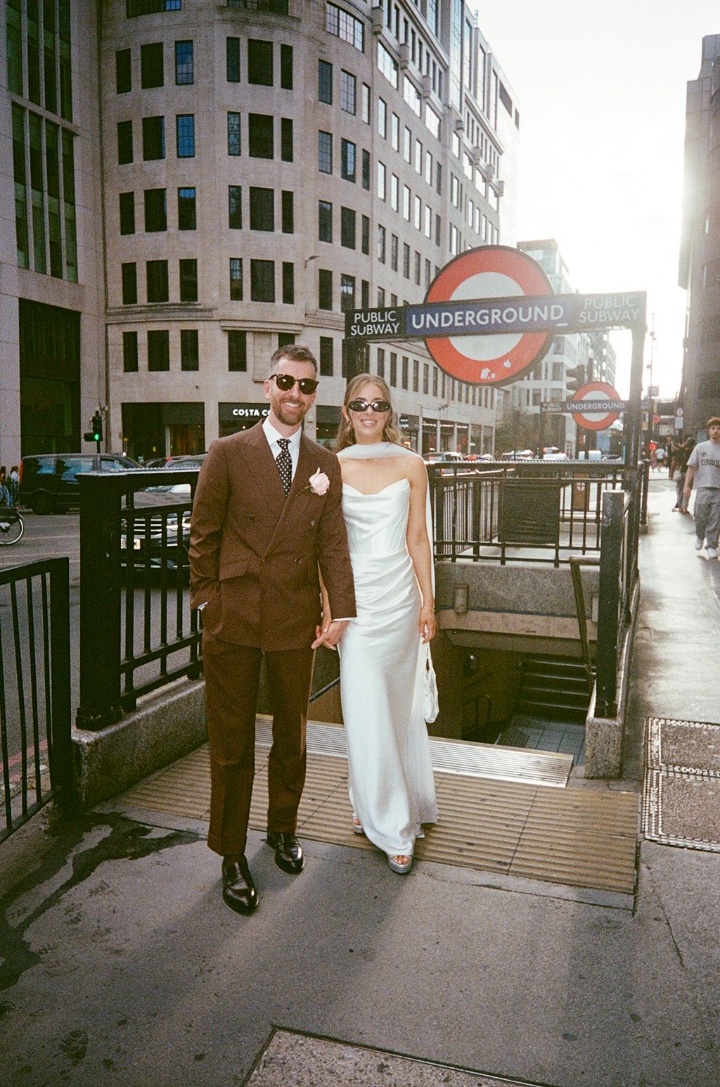 Wedding couple in white dress and brown suit posing at London Underground entrance.