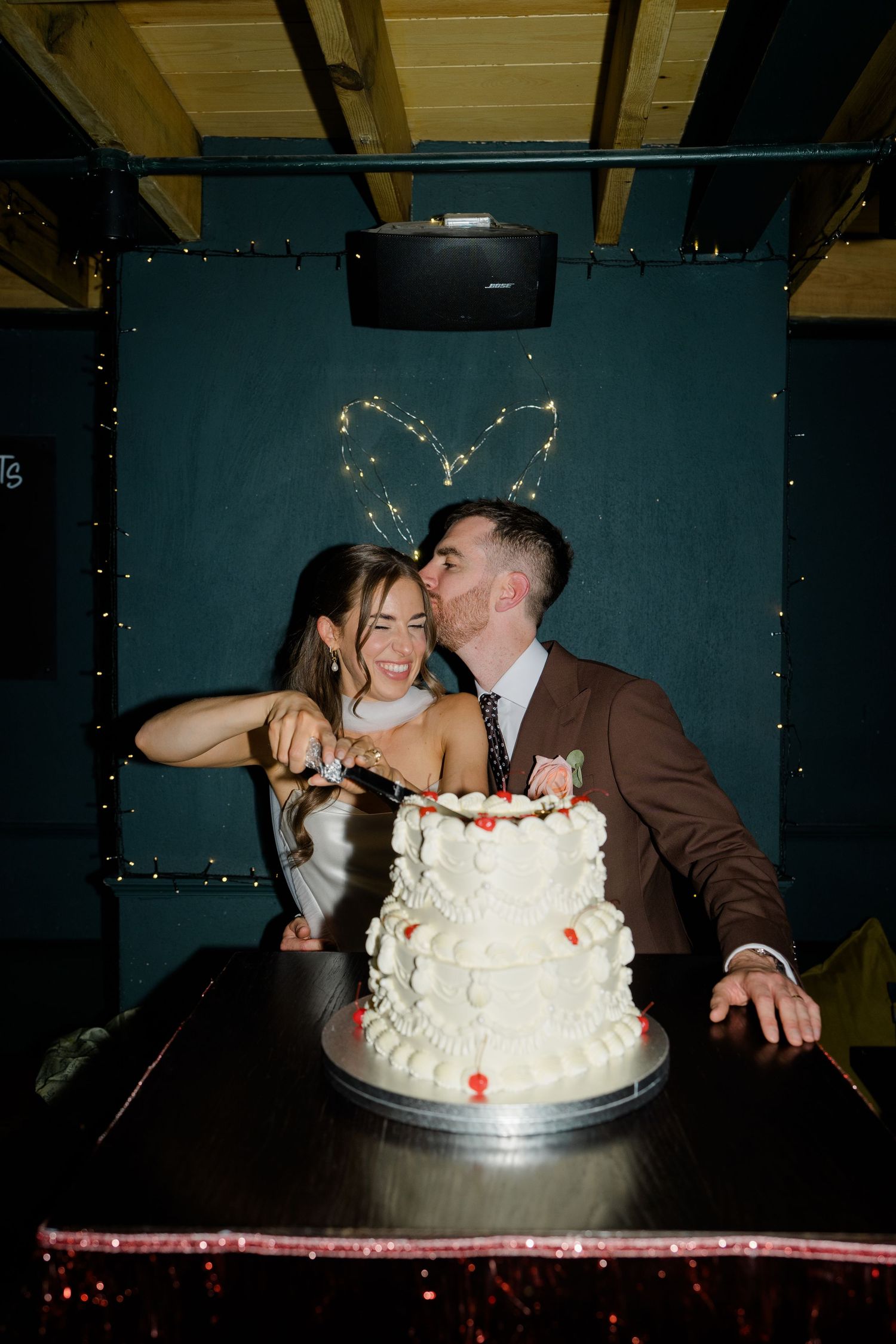 Bride and groom kissing while cutting tiered white wedding cake.