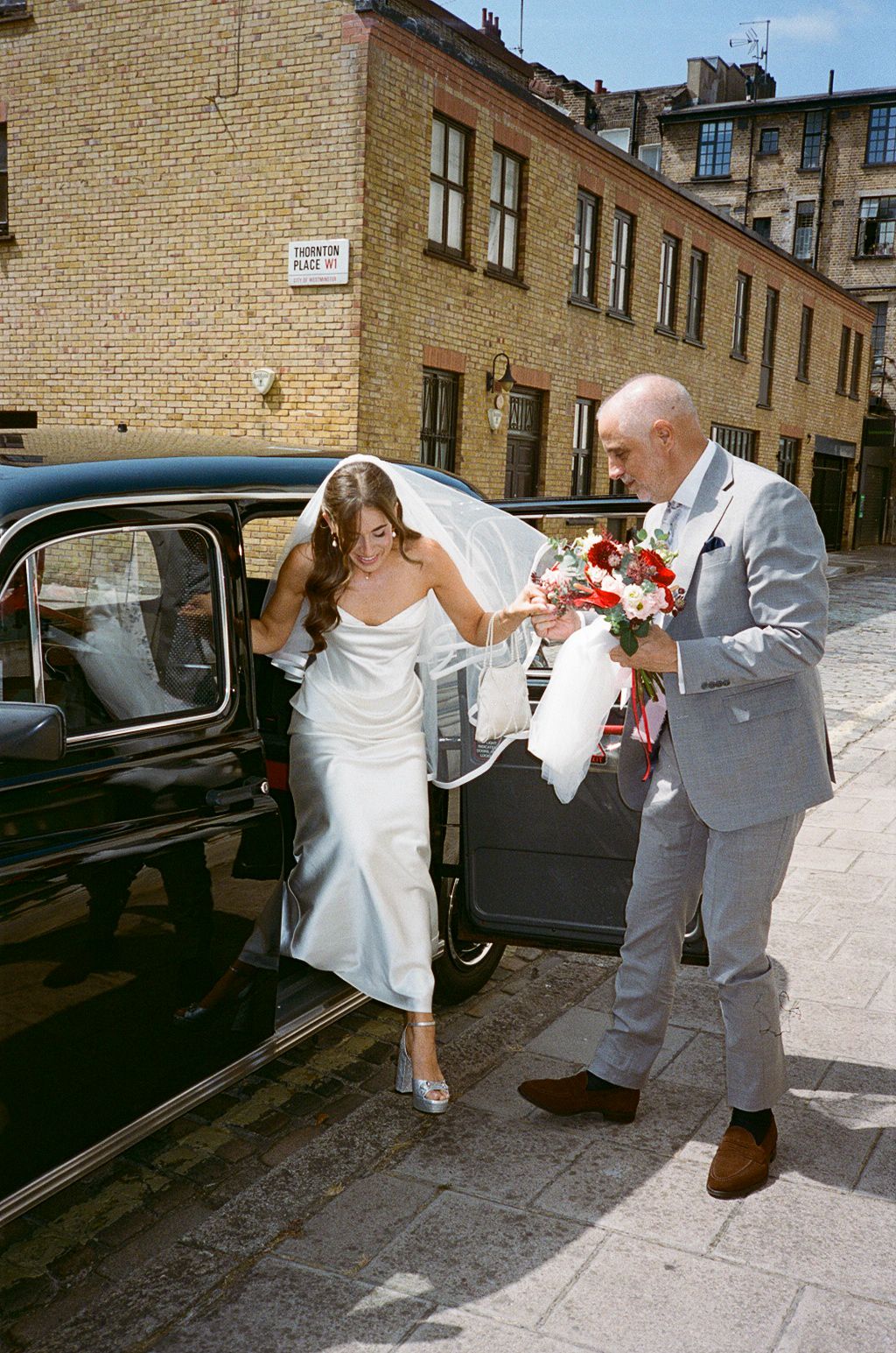 Bride in white midi satin dress and veil exits black vintage car assisted by man in grey suit holding bouquet.