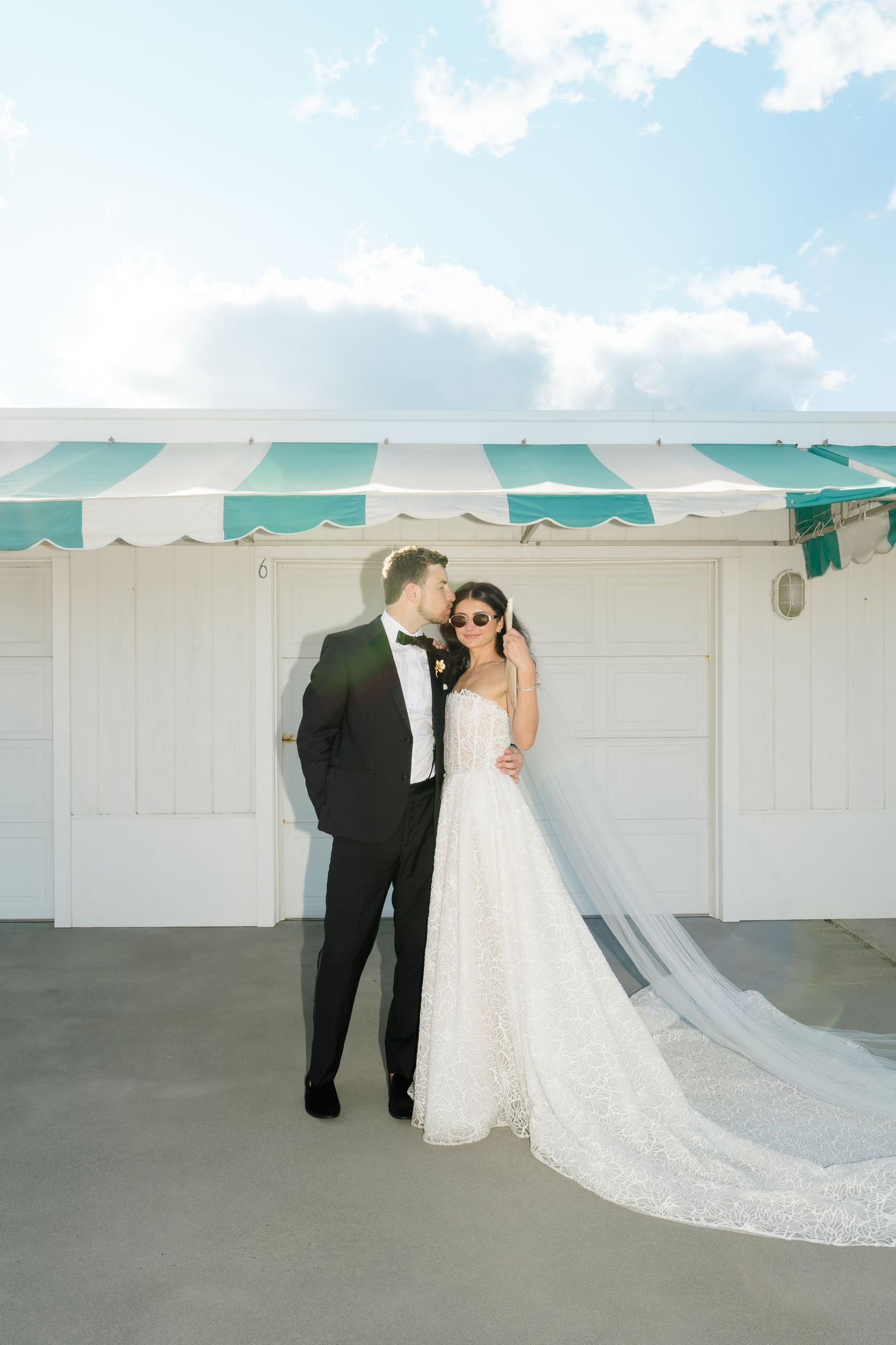 Bride and groom share a romantic kiss in front of a white building with teal striped awning on their wedding day.
