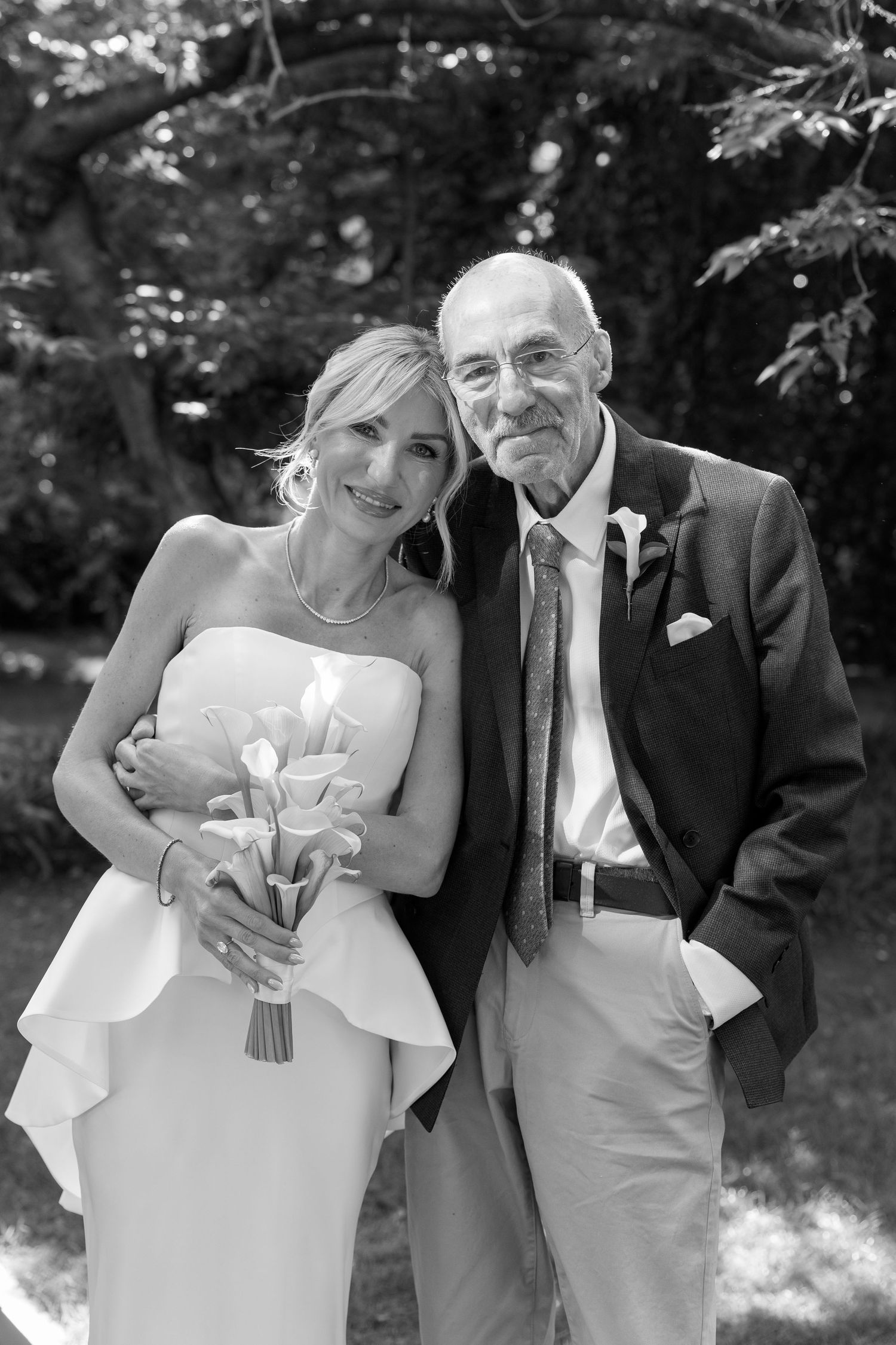 Black and white wedding photo of a smiling bride in white dress holding calla lilies beside an elderly gentleman in a suit.