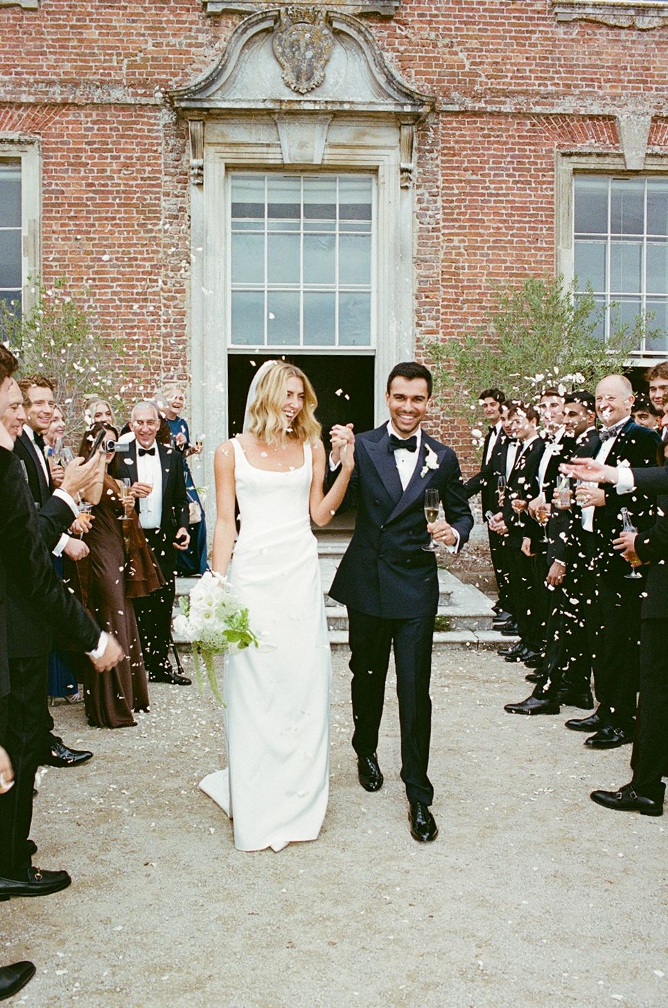 Bride and groom walk through confetti shower outside brick venue, she in white gown, he in navy tuxedo.
