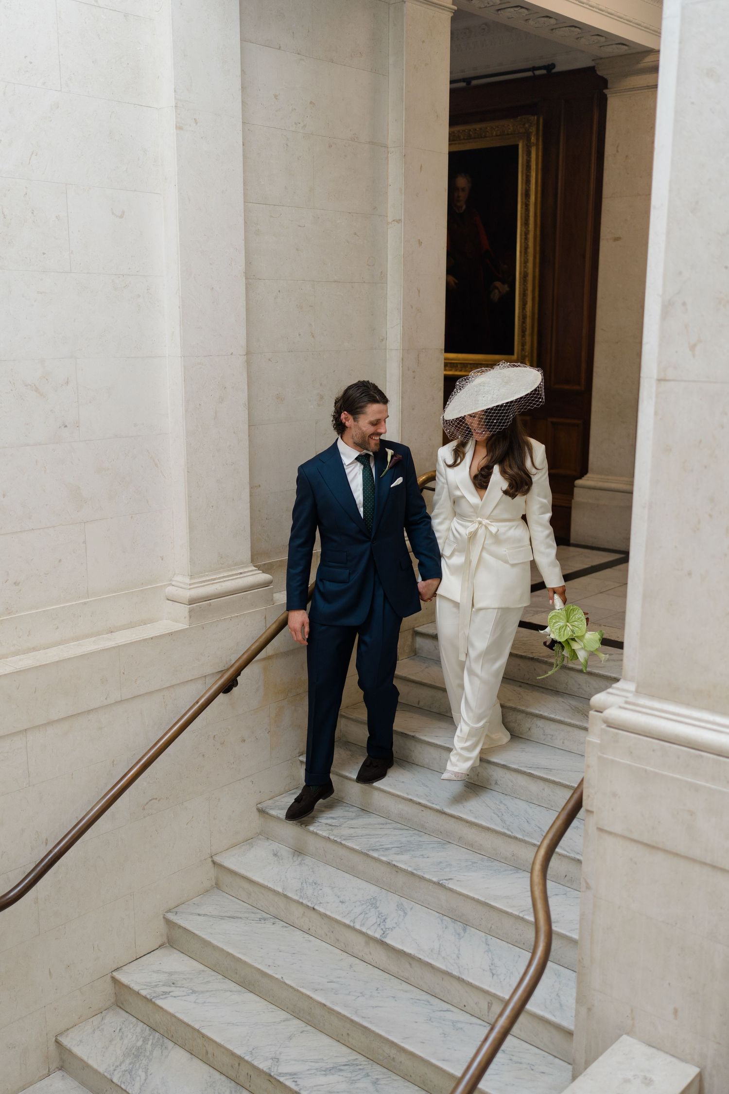 Bride and groom in white and navy suits walk down marble staircase inside elegant wedding venue.