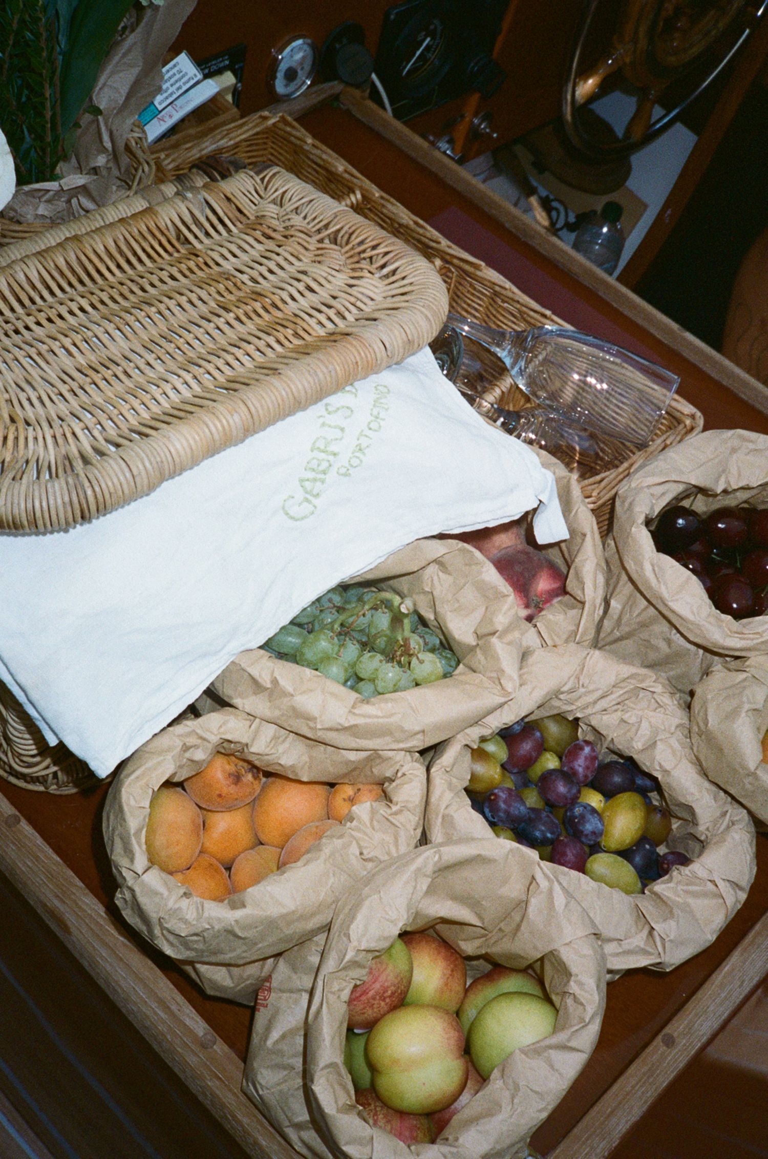 Assorted fresh fruits in brown paper bags on a wooden table with a wicker basket tray nearby.