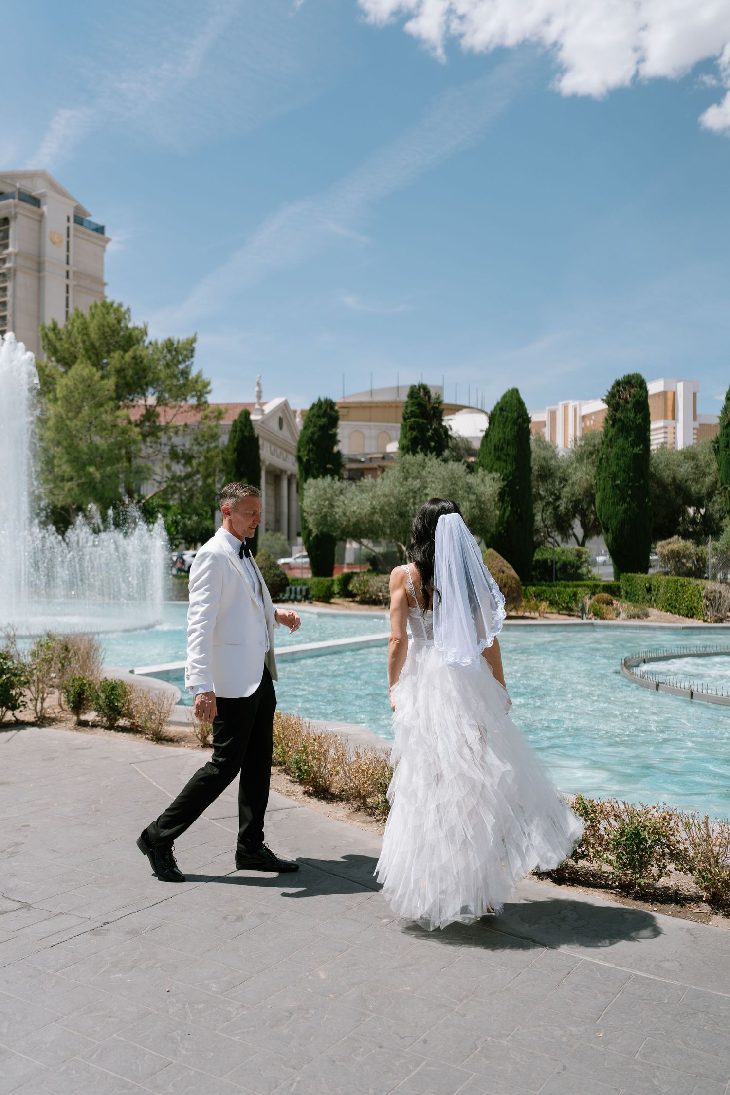 Wedding couple walks along resort pool area with fountains and Las Vegas skyline in background.