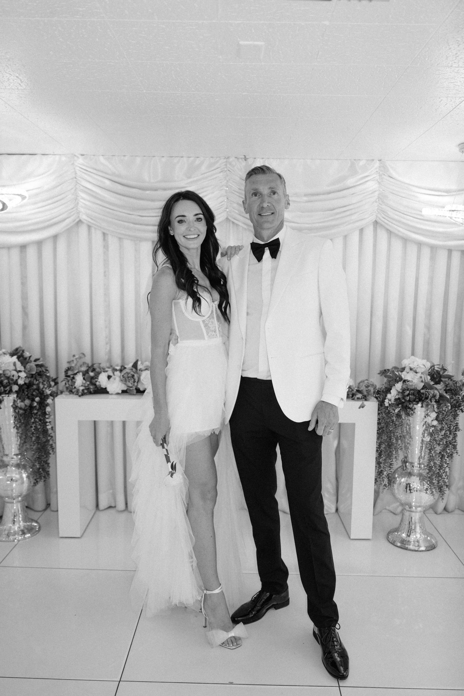 Formal wedding portrait in front of a white draped backdrop with floral arrangements.