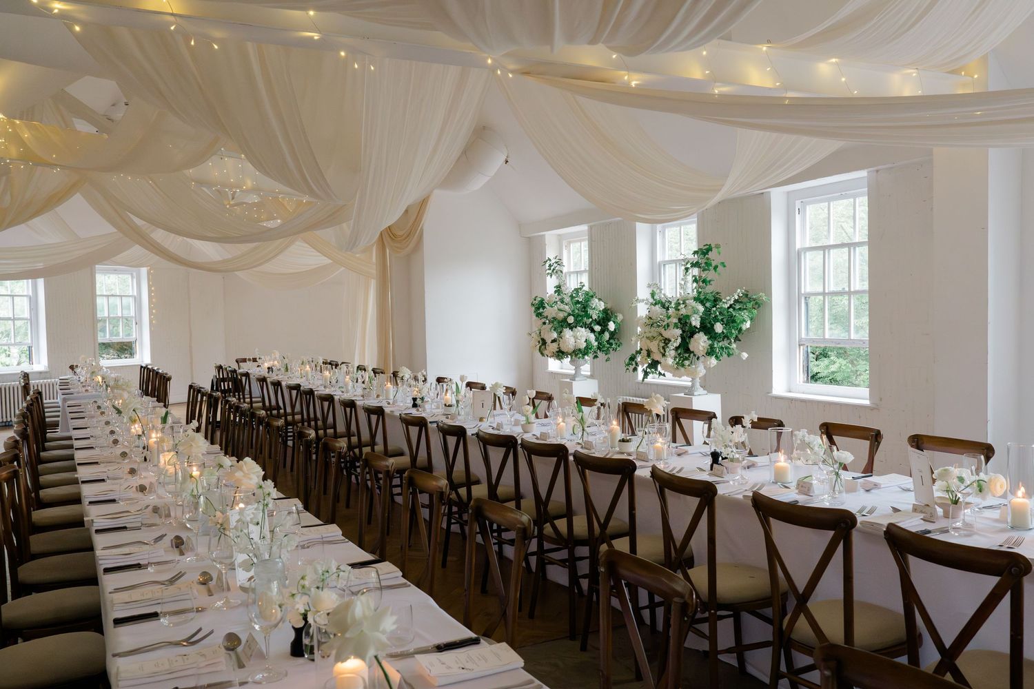 Elegant wedding reception hall with long tables, white draping, fairy lights, and floral centerpieces.