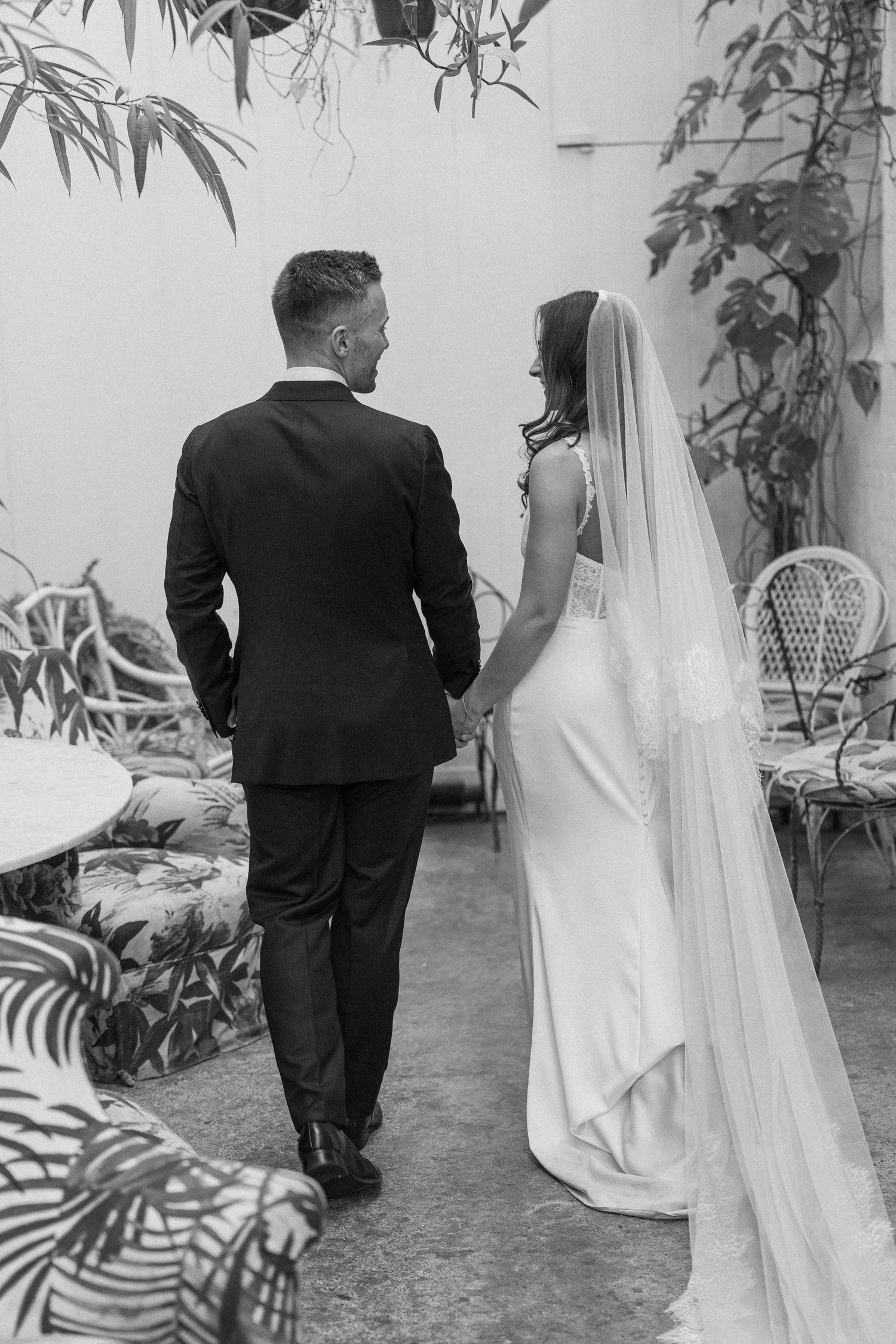 Bride and groom in black and white photo walk hand in hand through tropical garden venue.