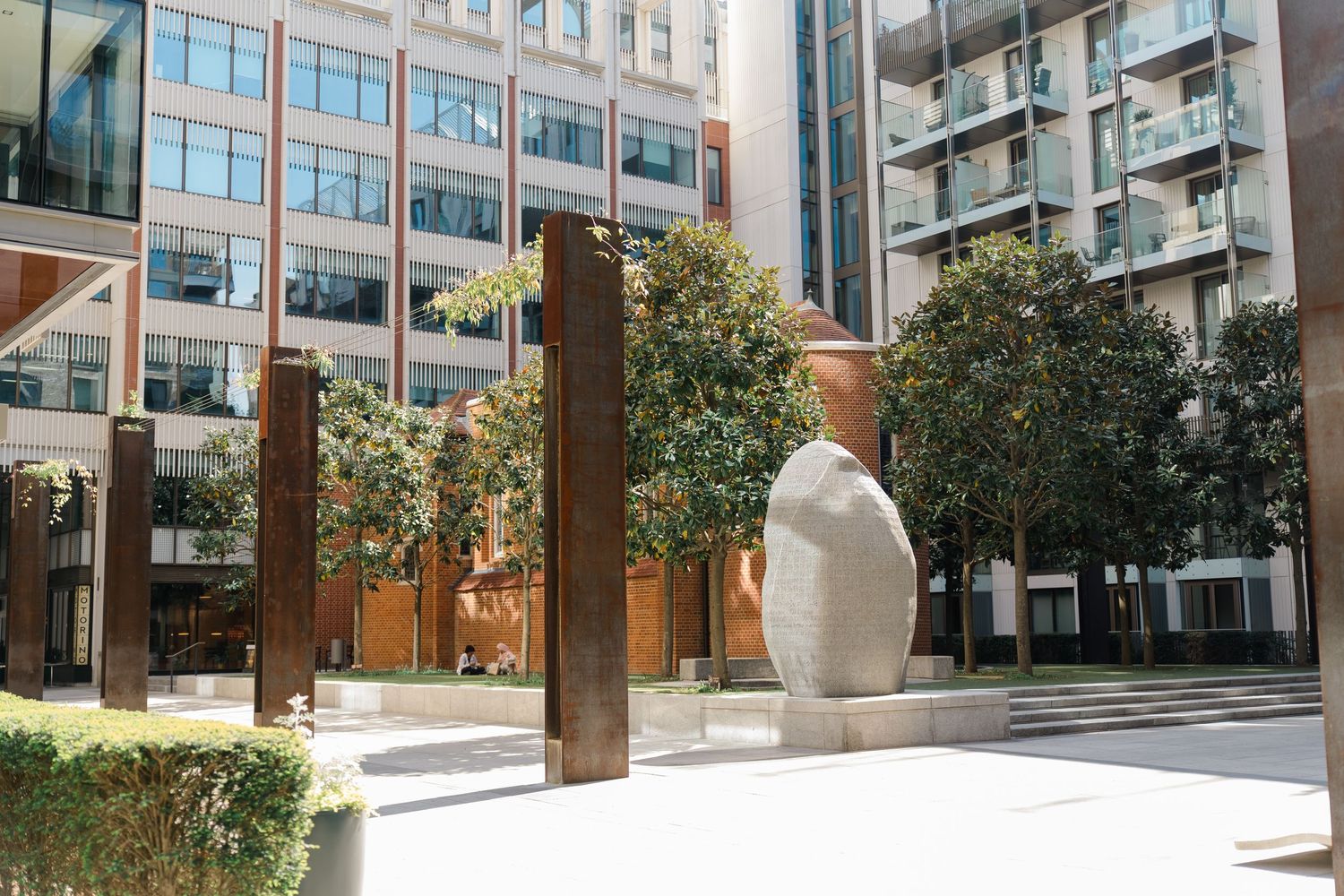 Modern urban courtyard with a large abstract stone sculpture and tall russet steel columns surrounded by lush green trees.