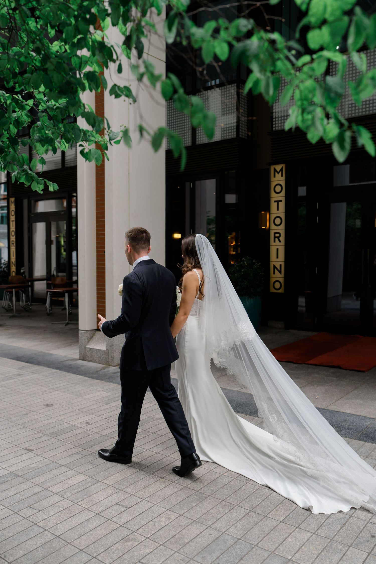 Bride and groom walking hand in hand outside Motorino restaurant, her long white veil trailing behind on city pavement.