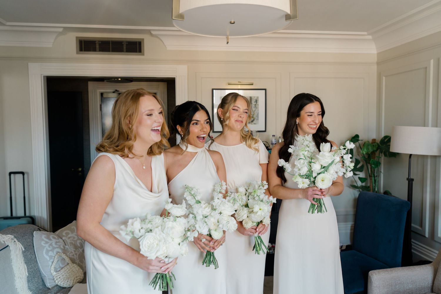 Four bridesmaids in white dresses holding white floral bouquets share a joyful moment in a bright hotel suite.