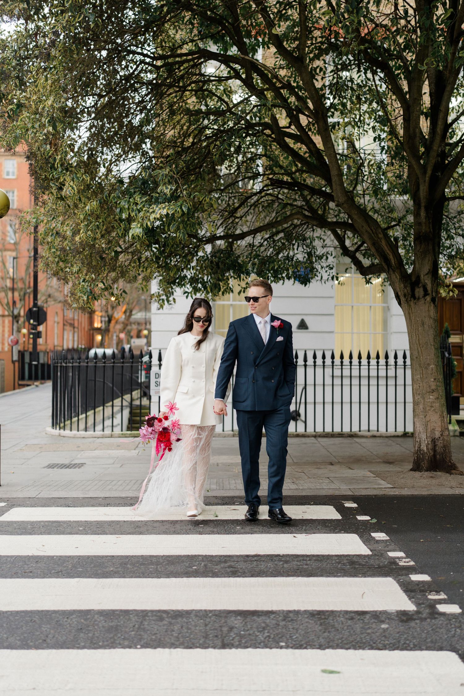 Bride and groom in sunglasses crossing zebra crossing on London street holding pink bouquet.