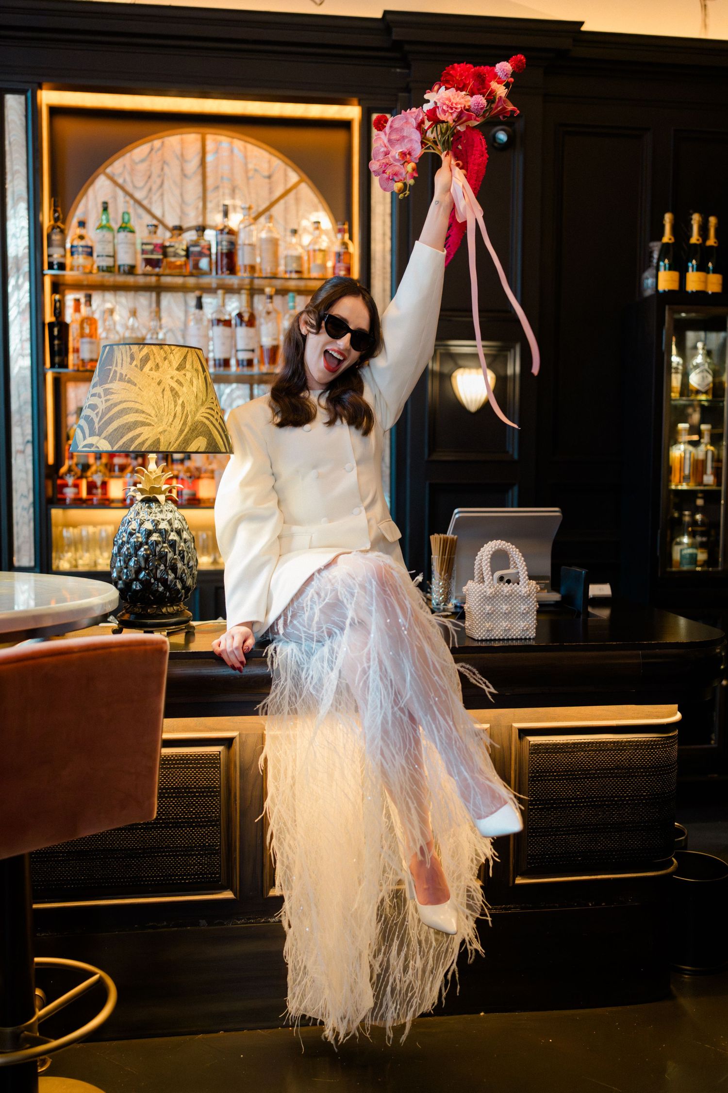 Bride in white feather skirt raises pink floral bouquet while sitting on bar counter at elegant venue.